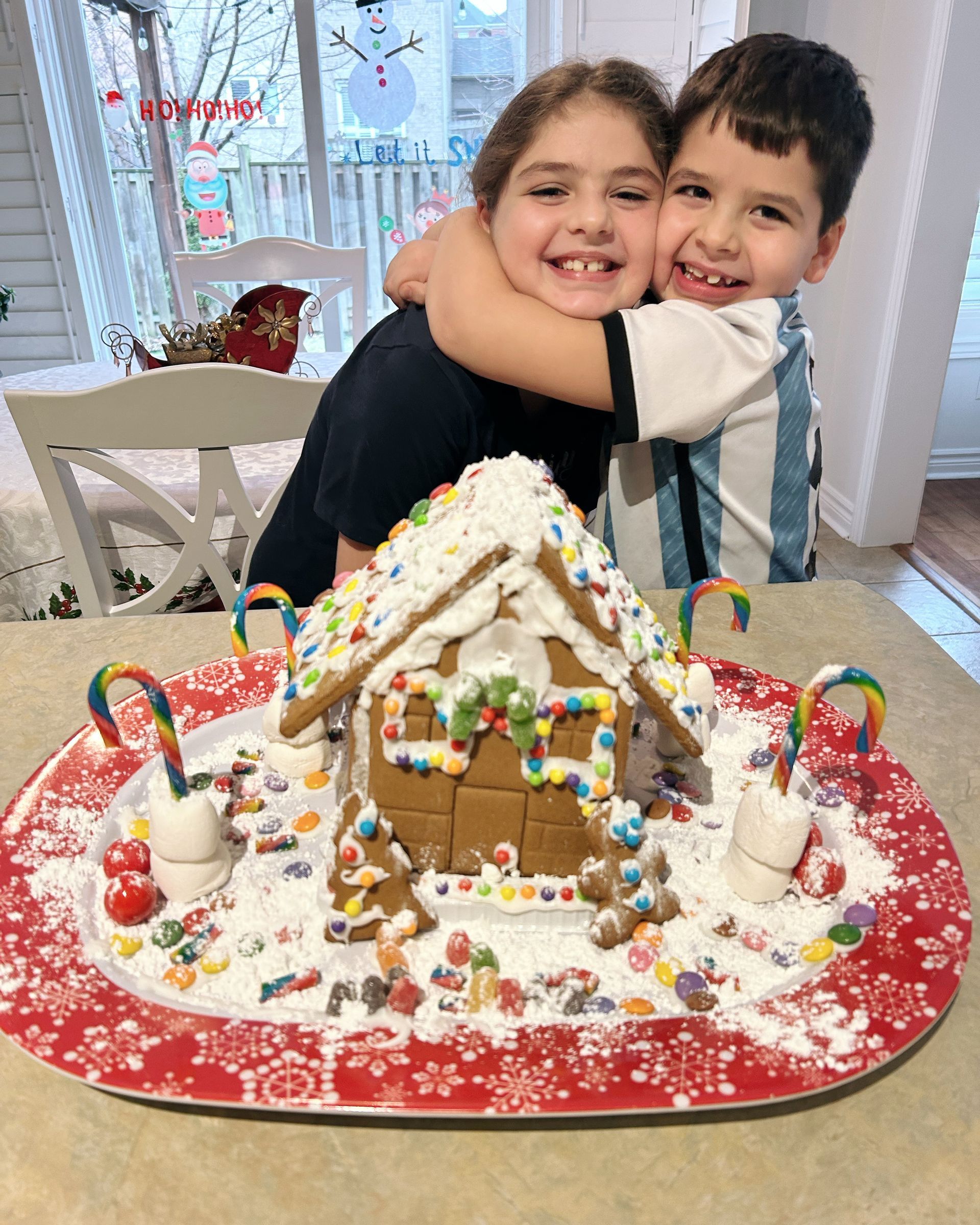 A boy and a girl are hugging in front of a gingerbread house.