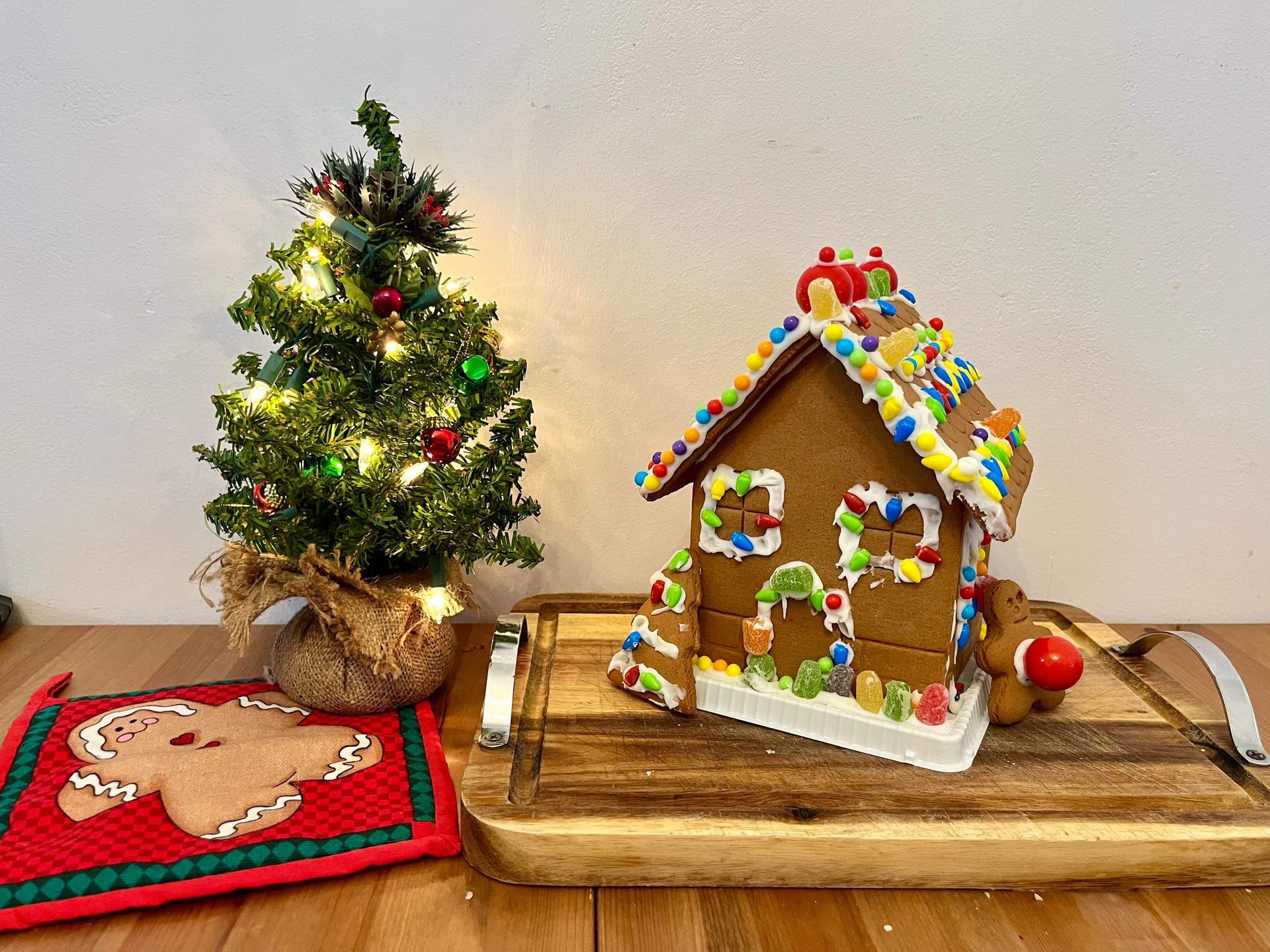 A gingerbread house is sitting on a wooden tray next to a small christmas tree.