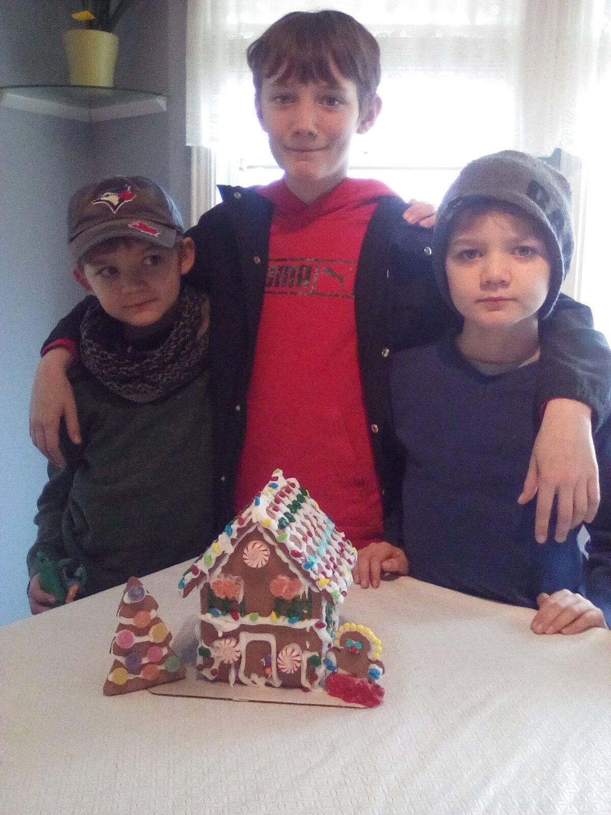 Three boys are standing next to a gingerbread house on a table.