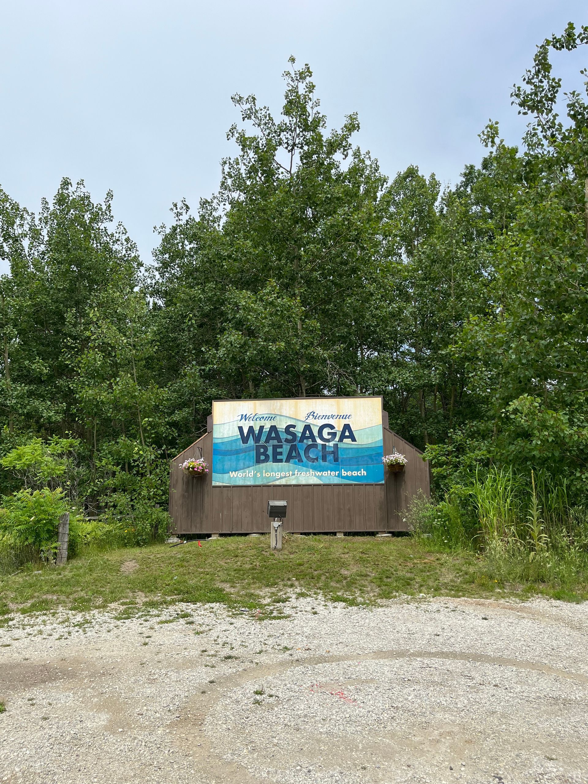 A sign for wasaga beach is surrounded by trees and gravel.