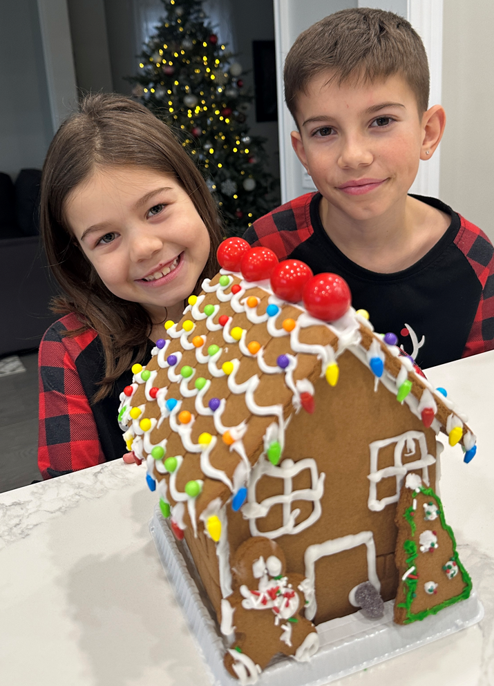 A boy and a girl are standing next to a gingerbread house.