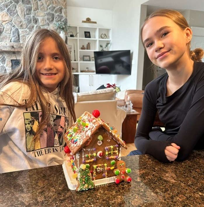 Two girls are sitting at a table with a gingerbread house on it