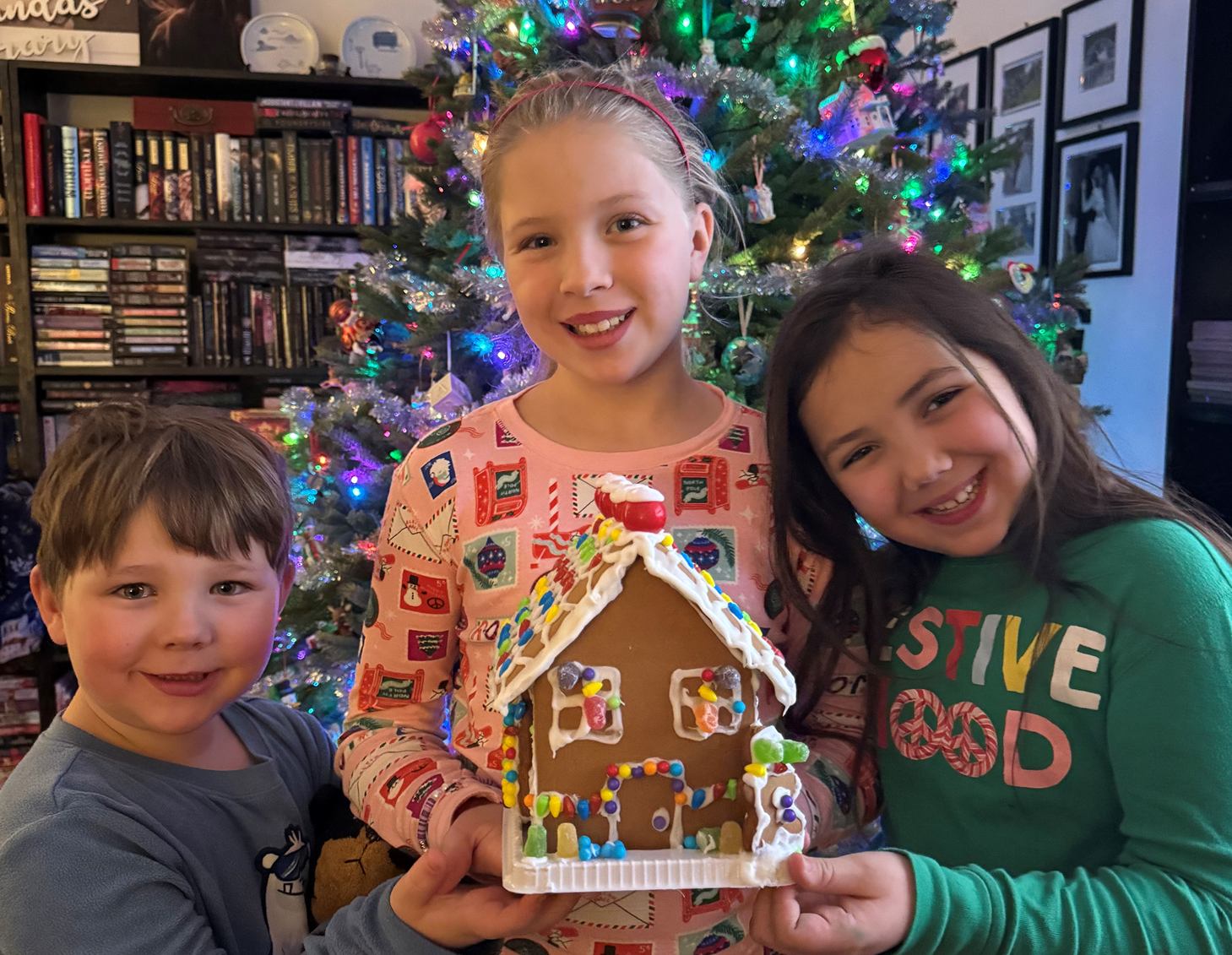 Three children are holding a gingerbread house in front of a christmas tree.