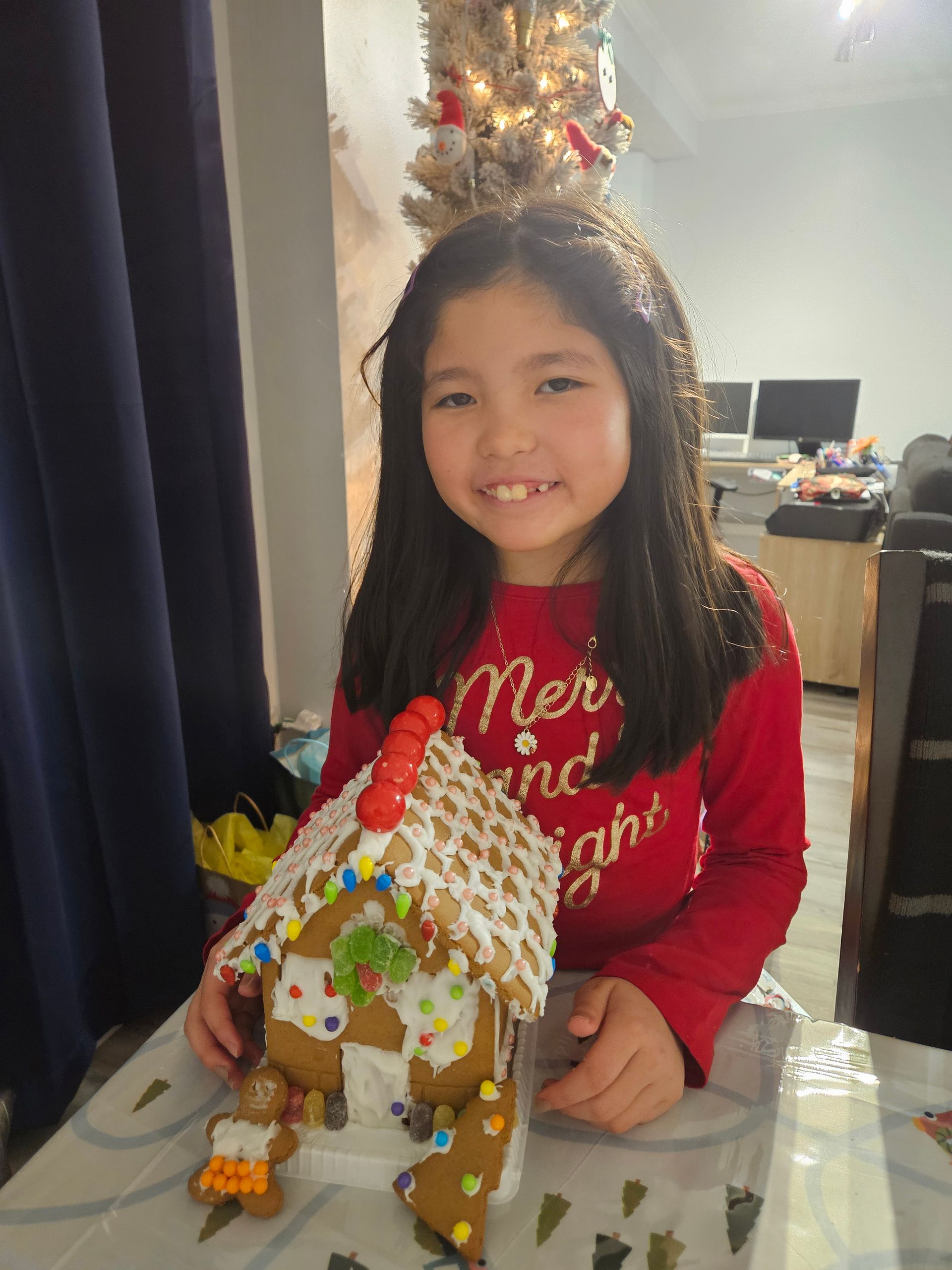 A little girl is sitting at a table holding a gingerbread house.