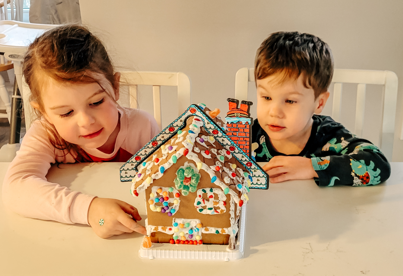A boy and a girl are sitting at a table looking at a gingerbread house.