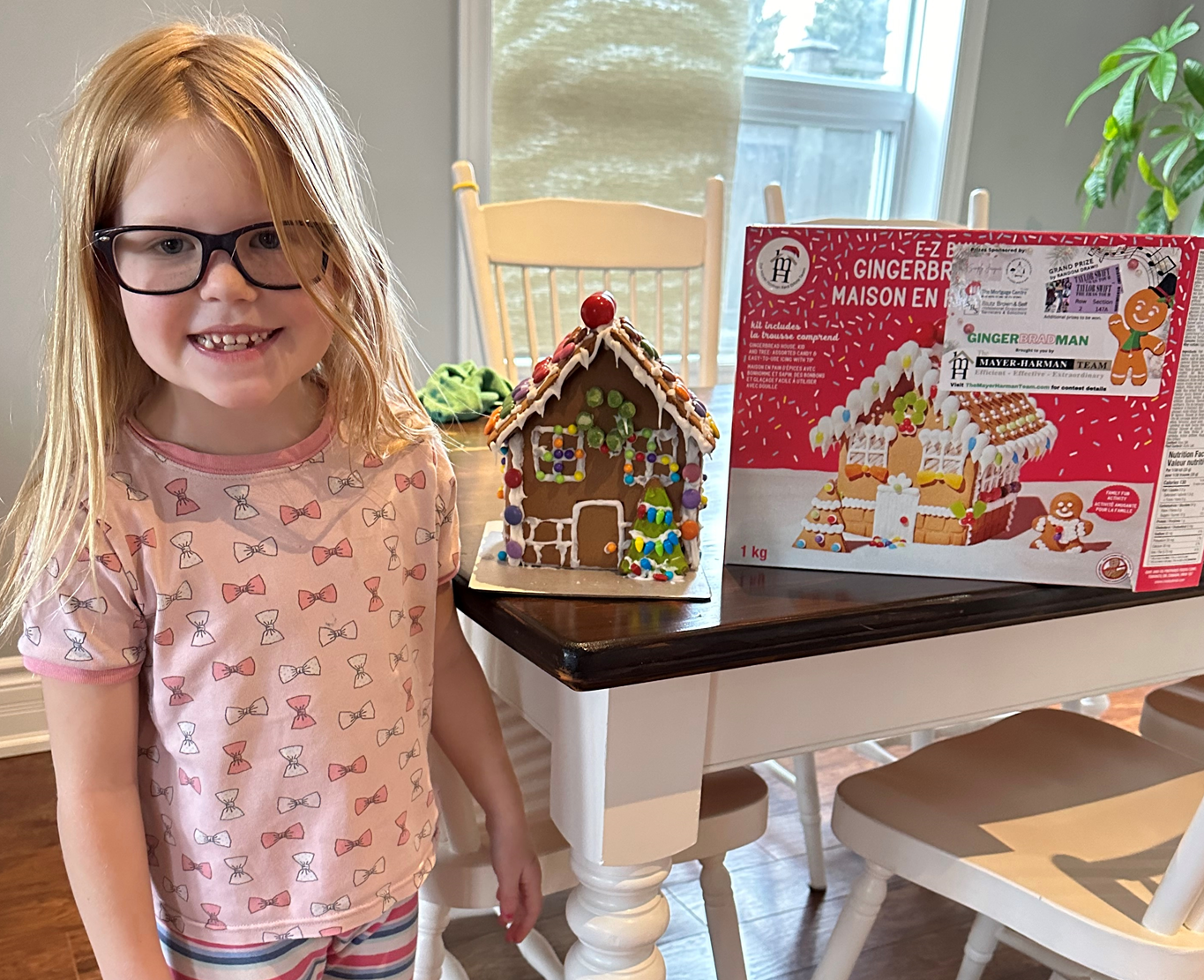 A little girl is standing next to a gingerbread house on a table.