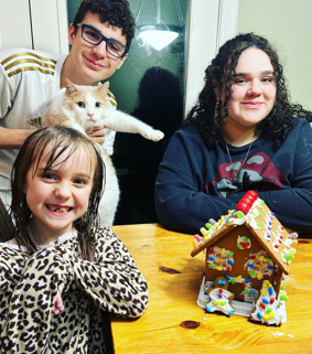 A group of people sitting at a table with a cat and a gingerbread house