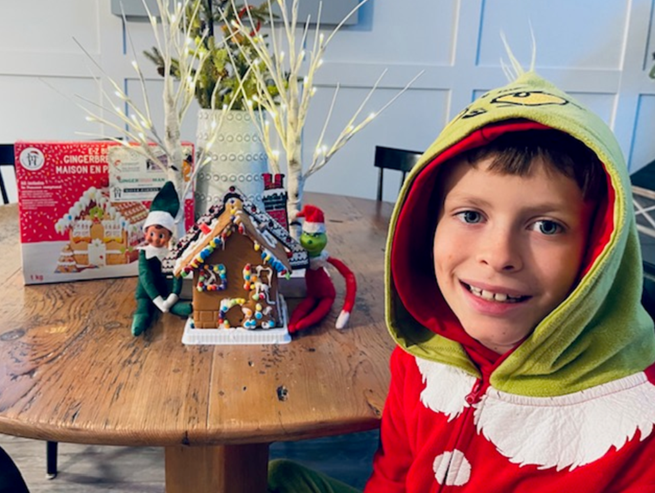 A young boy is sitting at a table with a gingerbread house and an elf on the shelf.