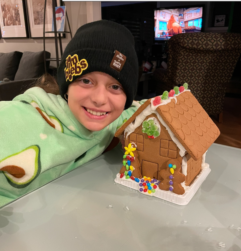 A young boy wearing a beanie is sitting next to a gingerbread house