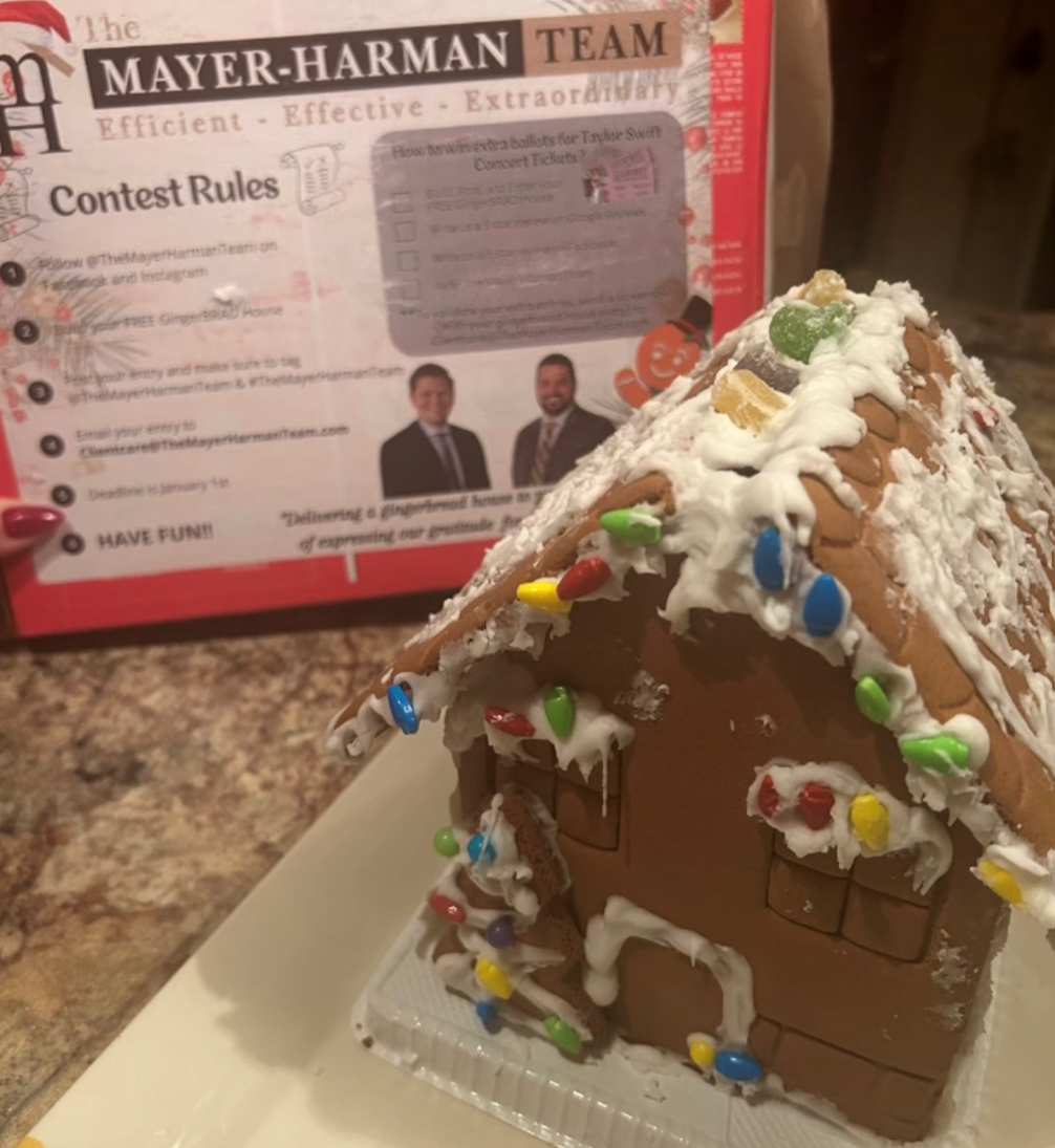 A gingerbread house sits on a table next to a mayer-harman team contest rules sign