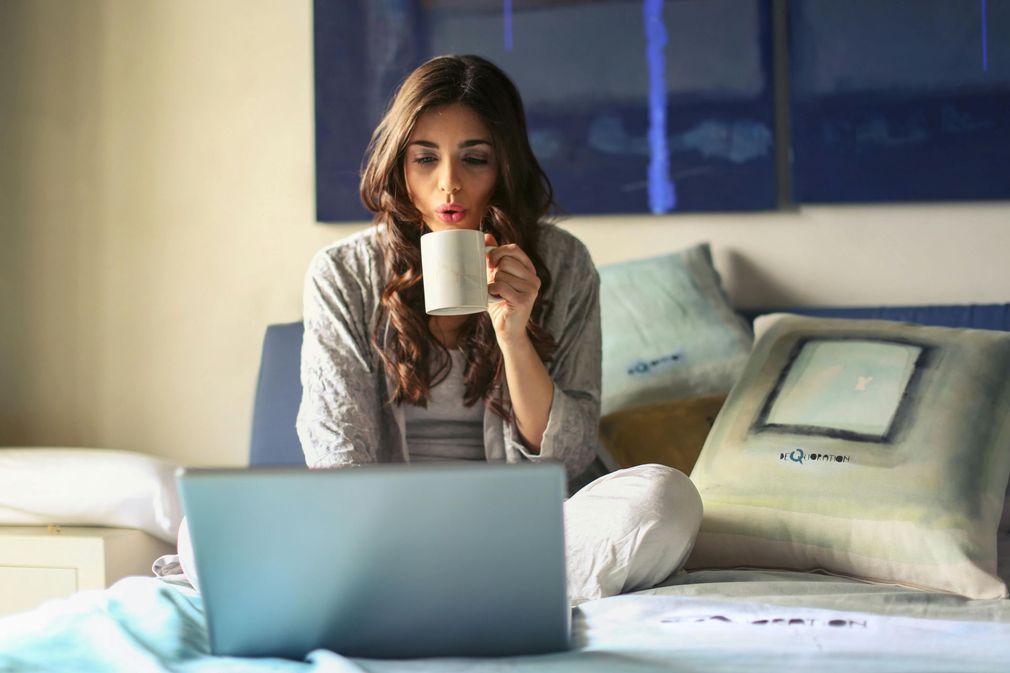 Woman drinking from mug while using a laptop, sitting on a bed, blue artwork in background.