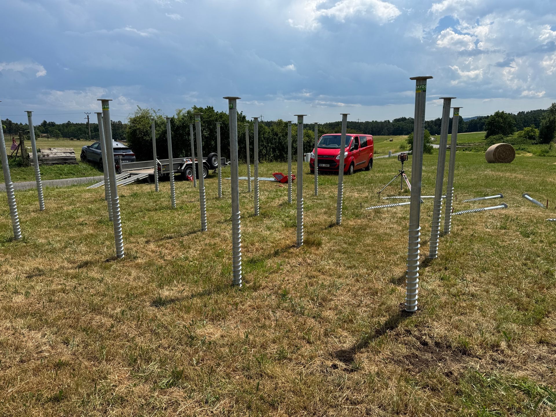 Metal ground screws installed in a grassy field, with a red van and other construction materials in the background.