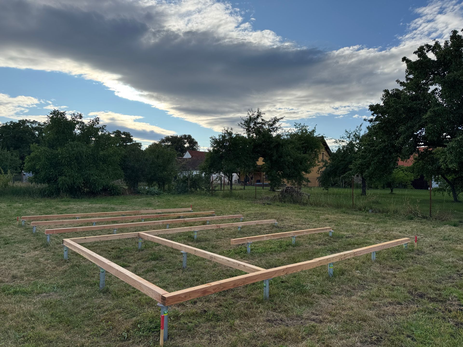Wooden frame foundation on metal posts set in grassy field, trees and cloudy sky background.
