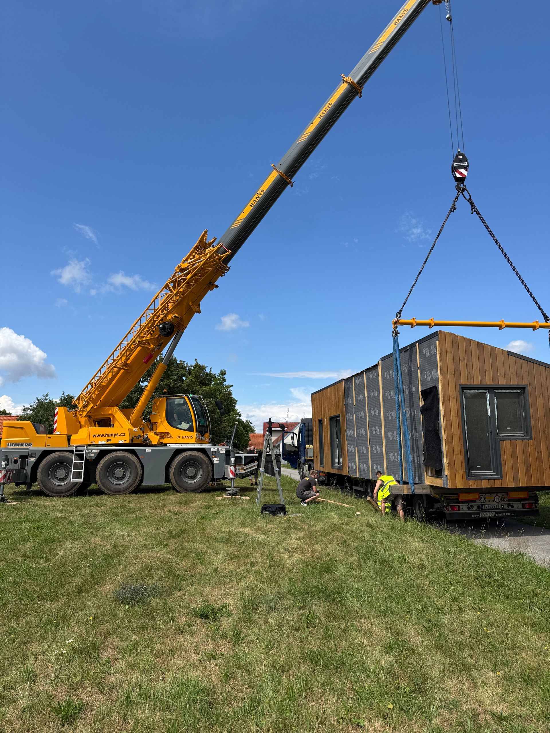 Yellow crane lifting a modular building section over a grassy area against a blue sky.