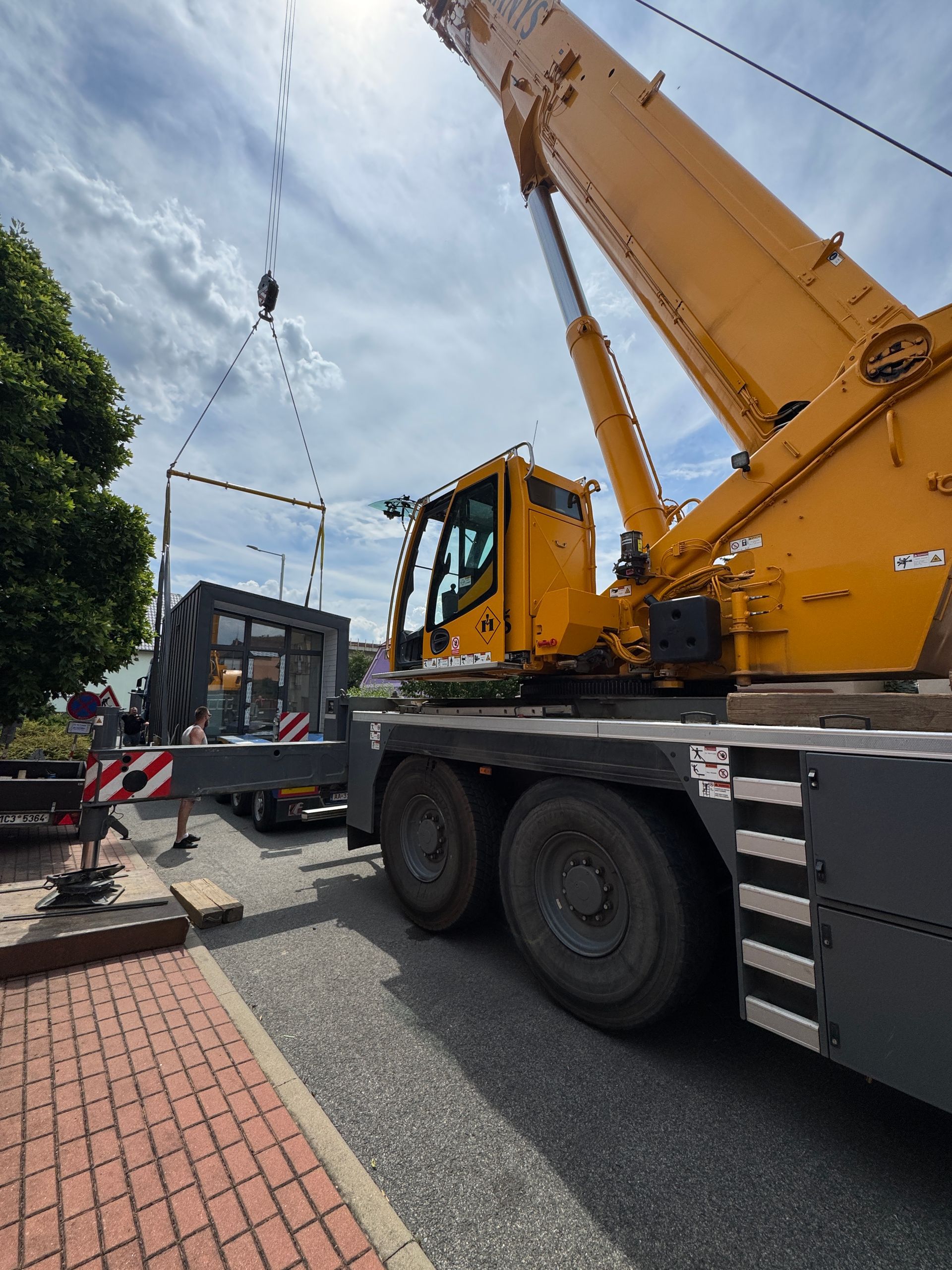 Yellow crane lifting a dark object on a city street; brick sidewalk in foreground, blue sky above.