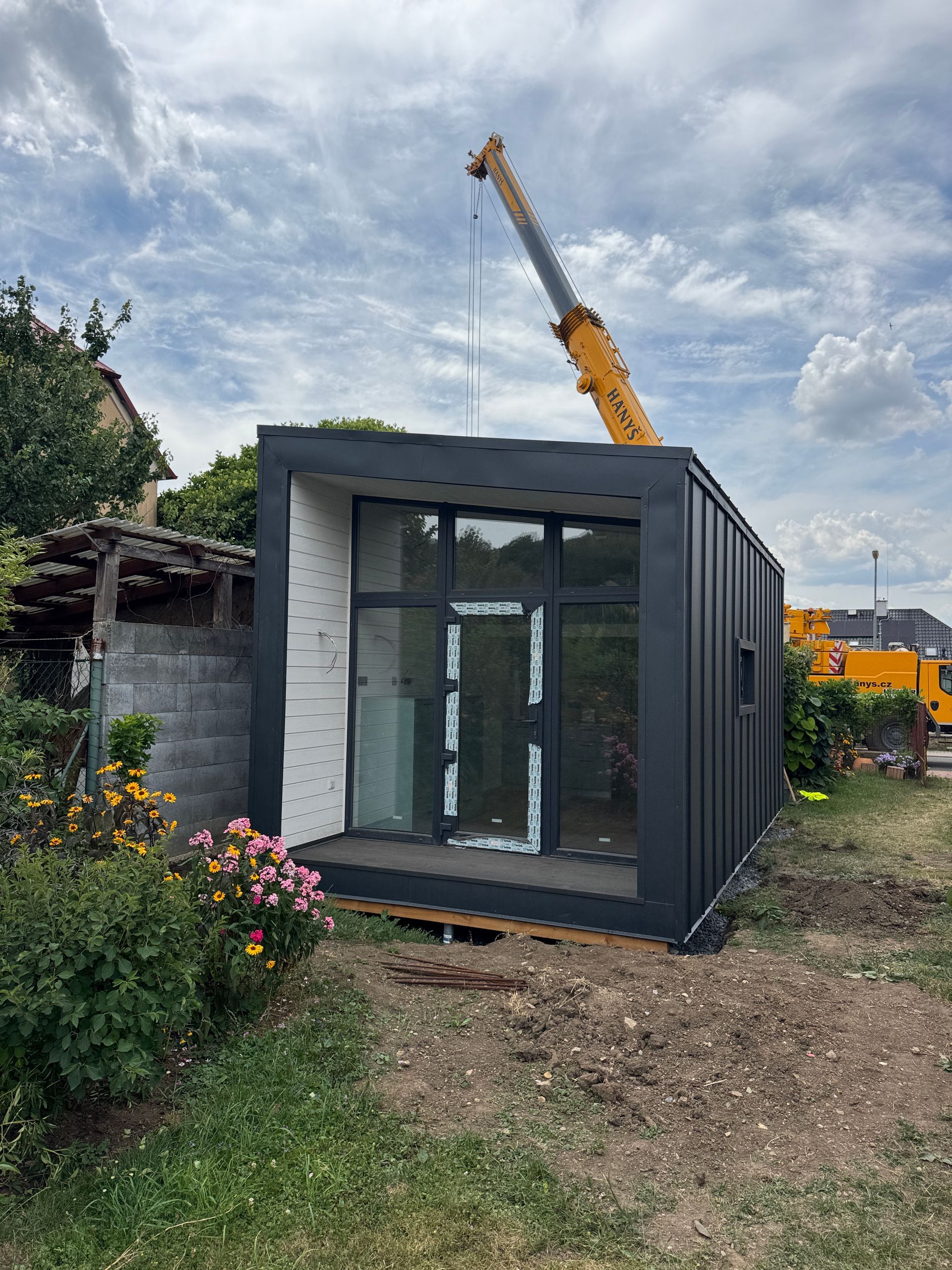 A modern dark gray modular building being set in place by a crane in a yard on a cloudy day.