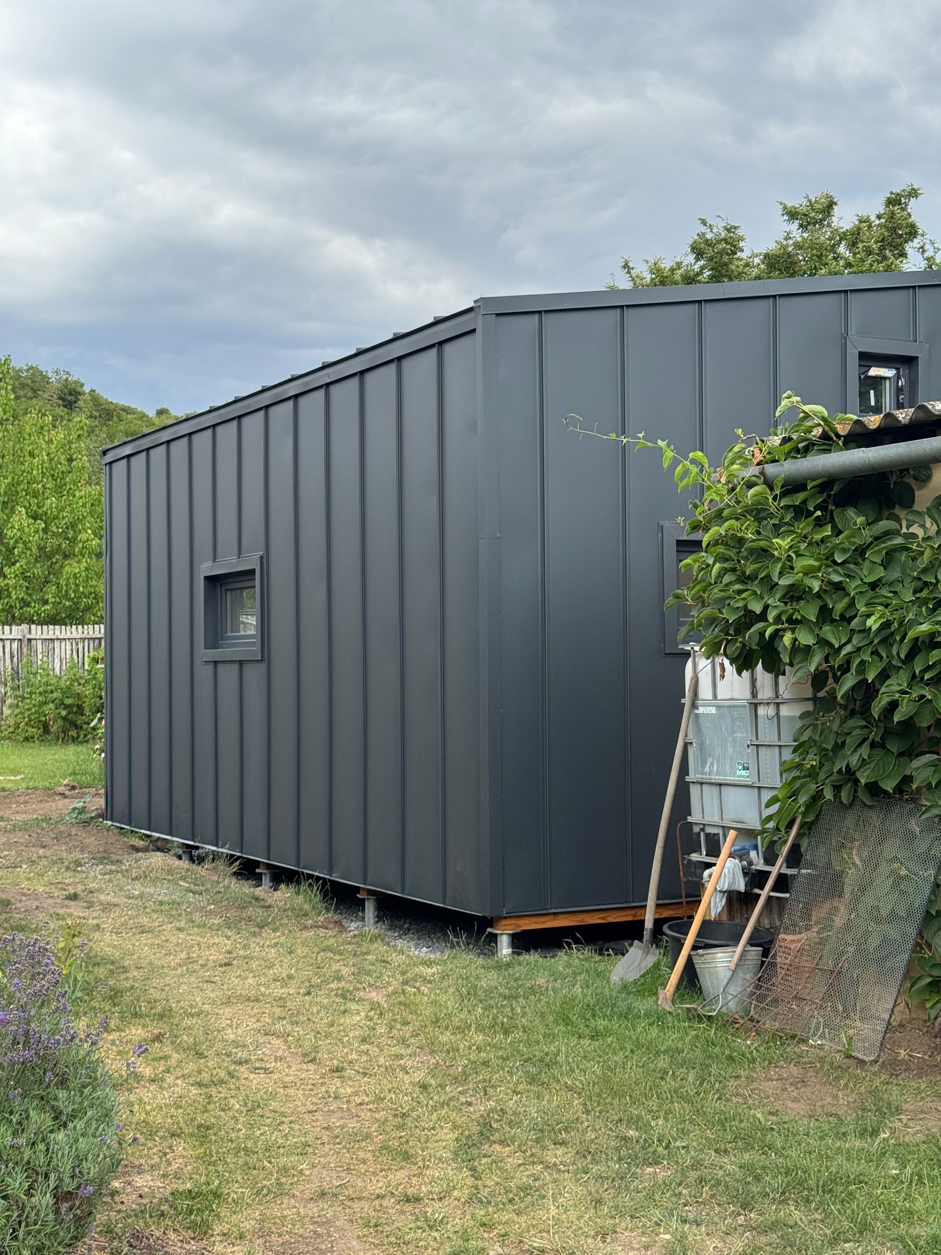 Dark gray, modern cabin with vertical siding sits in a grassy yard, surrounded by greenery.