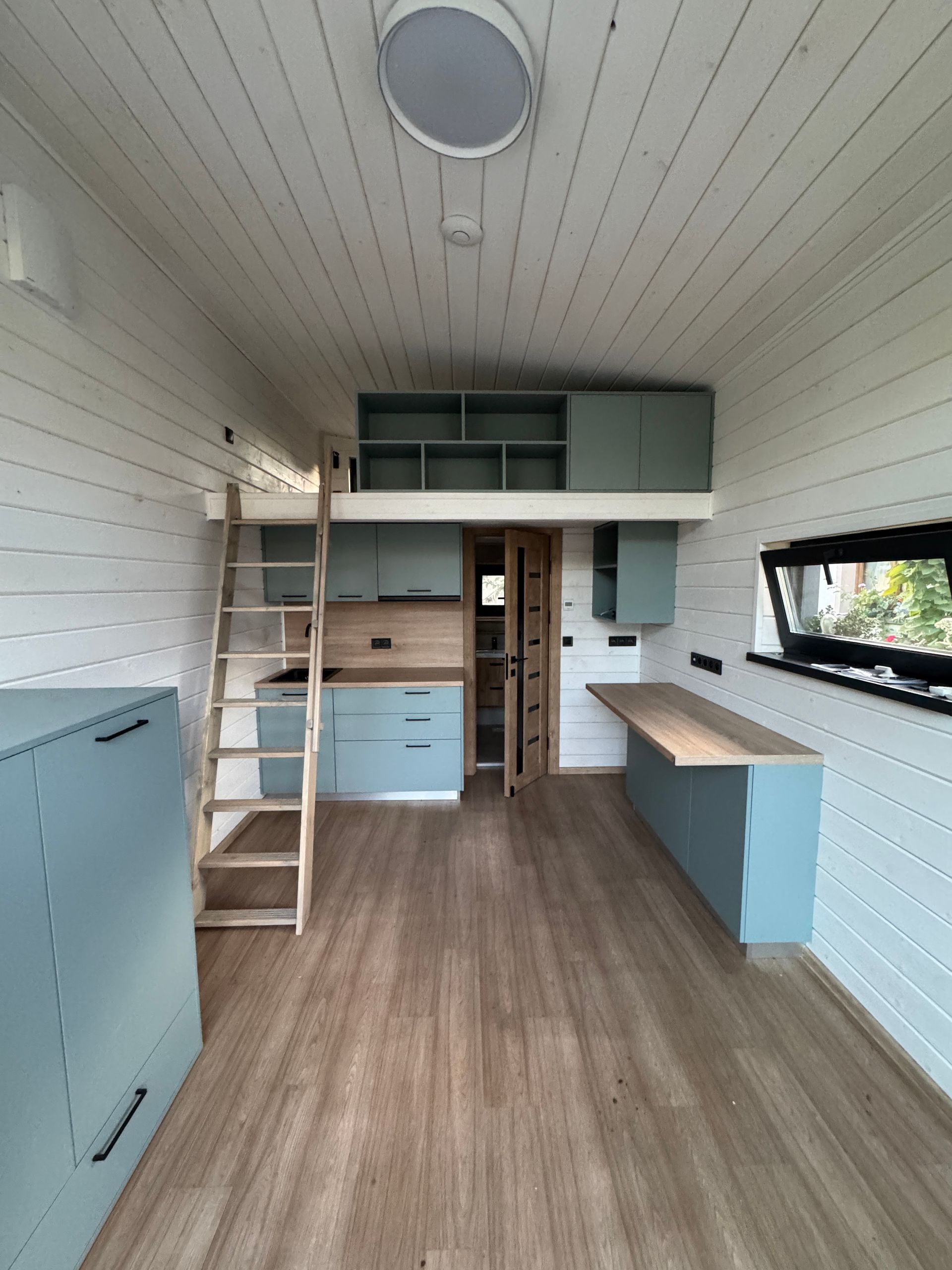Interior of a tiny home with a loft, light blue cabinetry, wood floors, and a skylight.