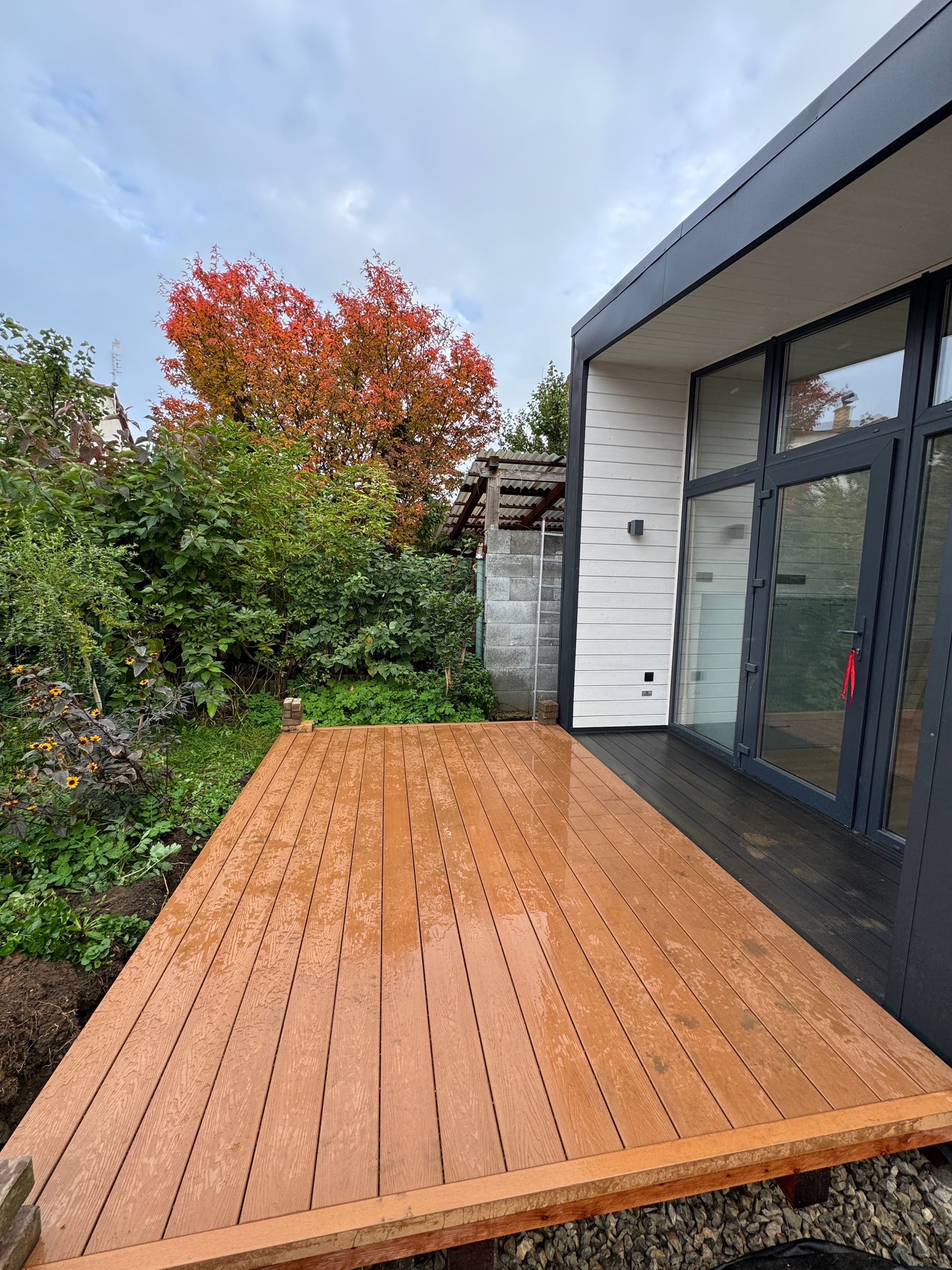 Wooden deck next to a modern building with glass doors, fall foliage in the background.