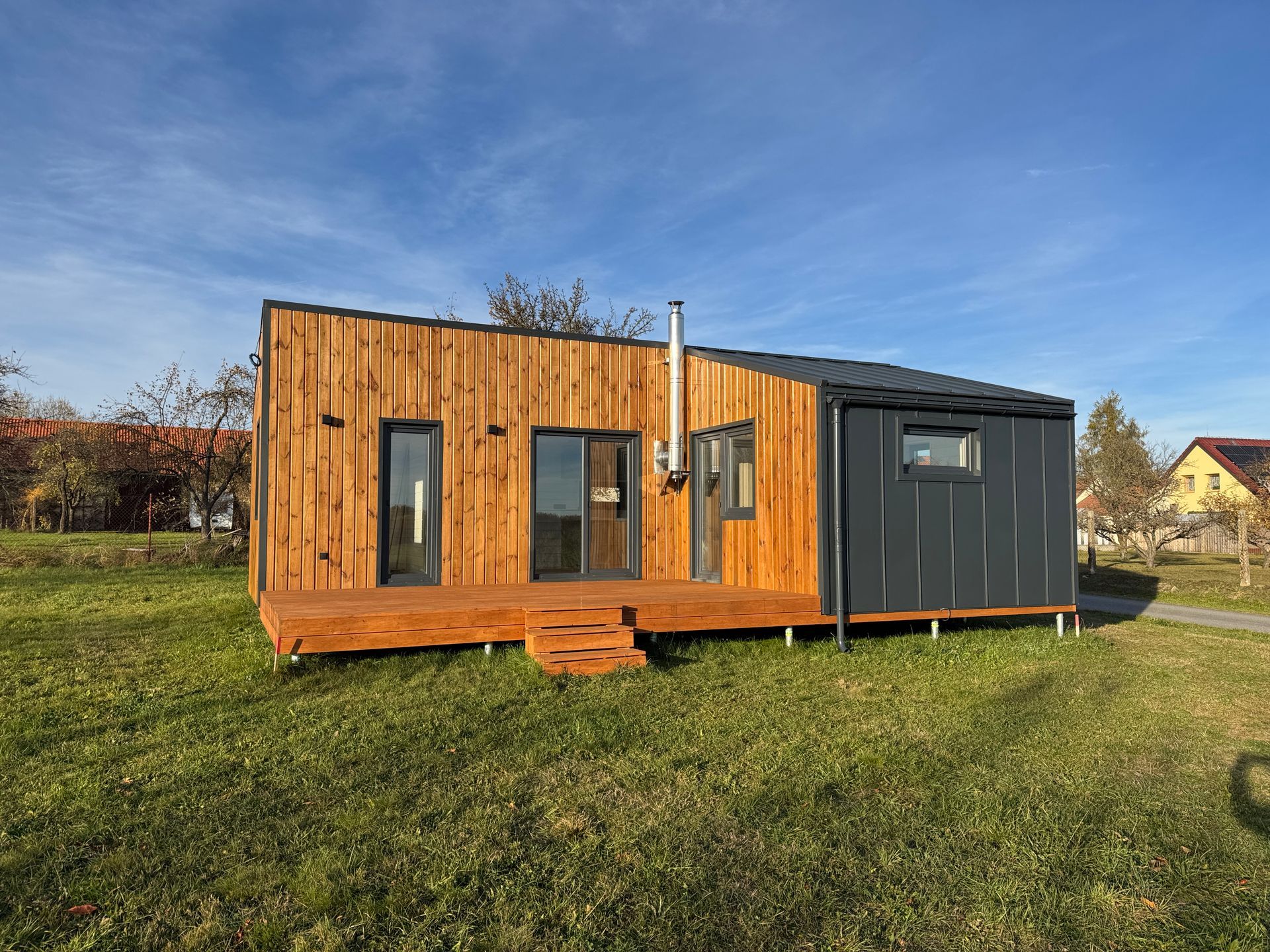 Wooden cabin with gray metal siding, deck, and chimney on a grassy field under a blue sky.