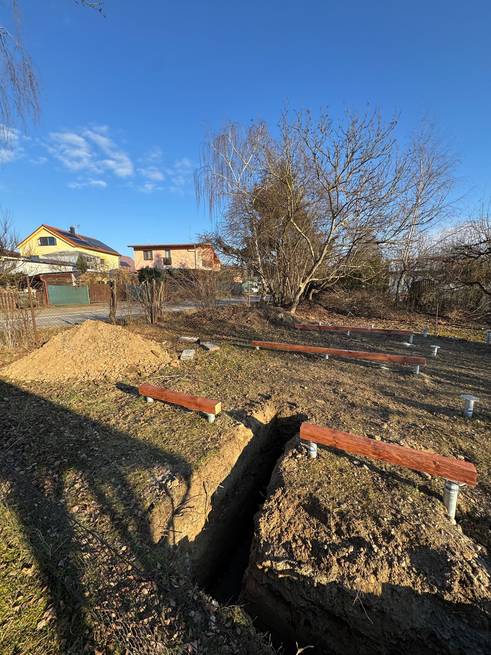 A construction site: trenches dug, wooden beams on metal posts, residential background, sunny day.