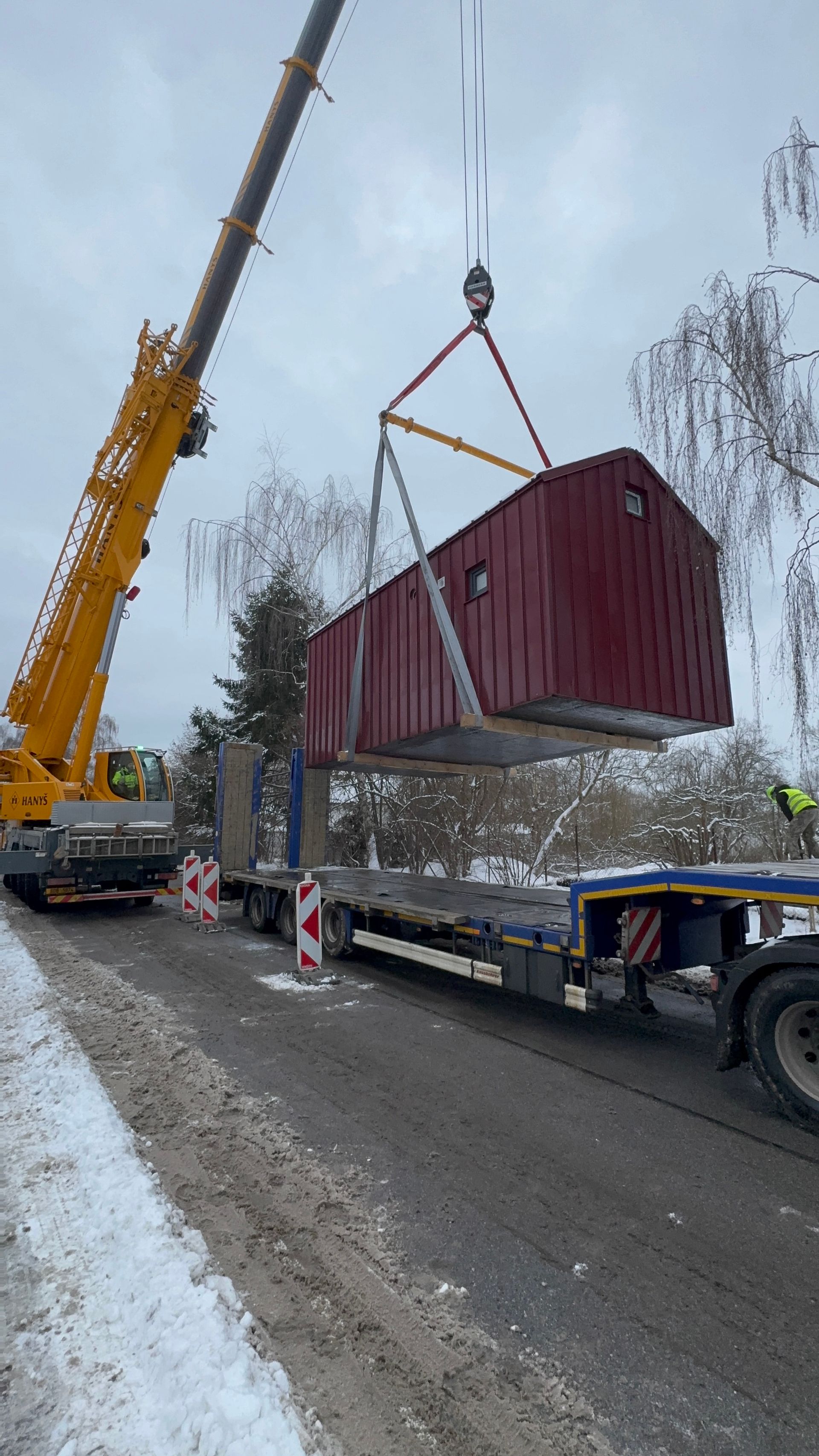 A crane lifts a maroon rectangular structure from a flatbed truck on a snowy street.