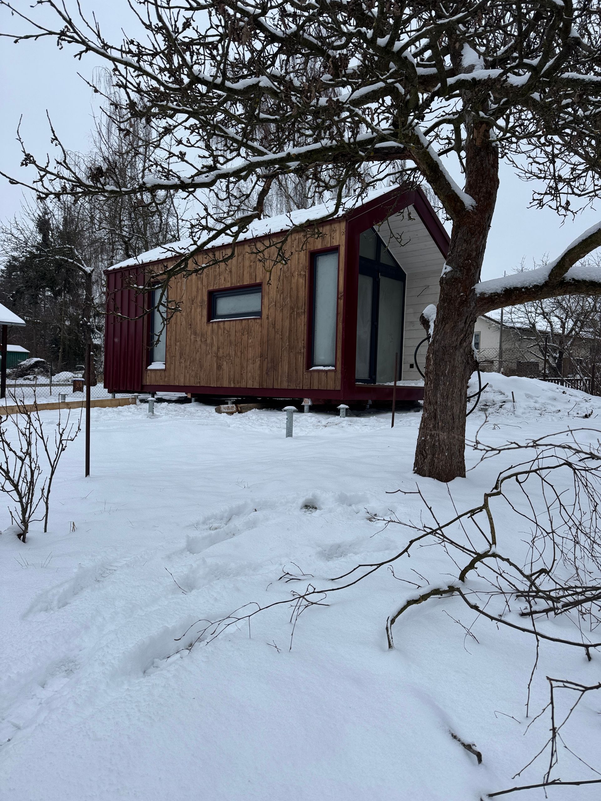 Tiny cabin in a snowy landscape. Brown and red exterior, with a tree in the foreground.