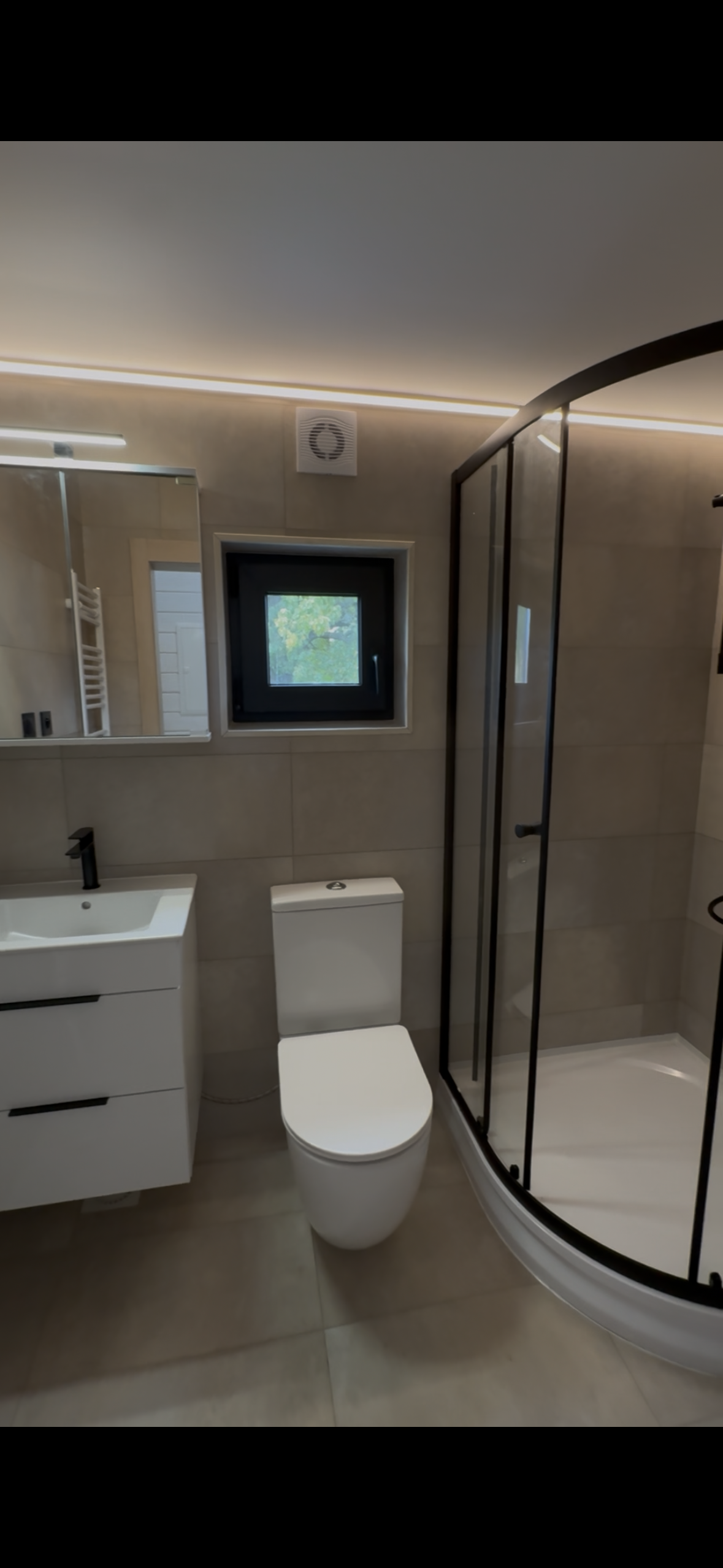 Modern bathroom with a white toilet, sink, and shower. Beige walls and black trim.
