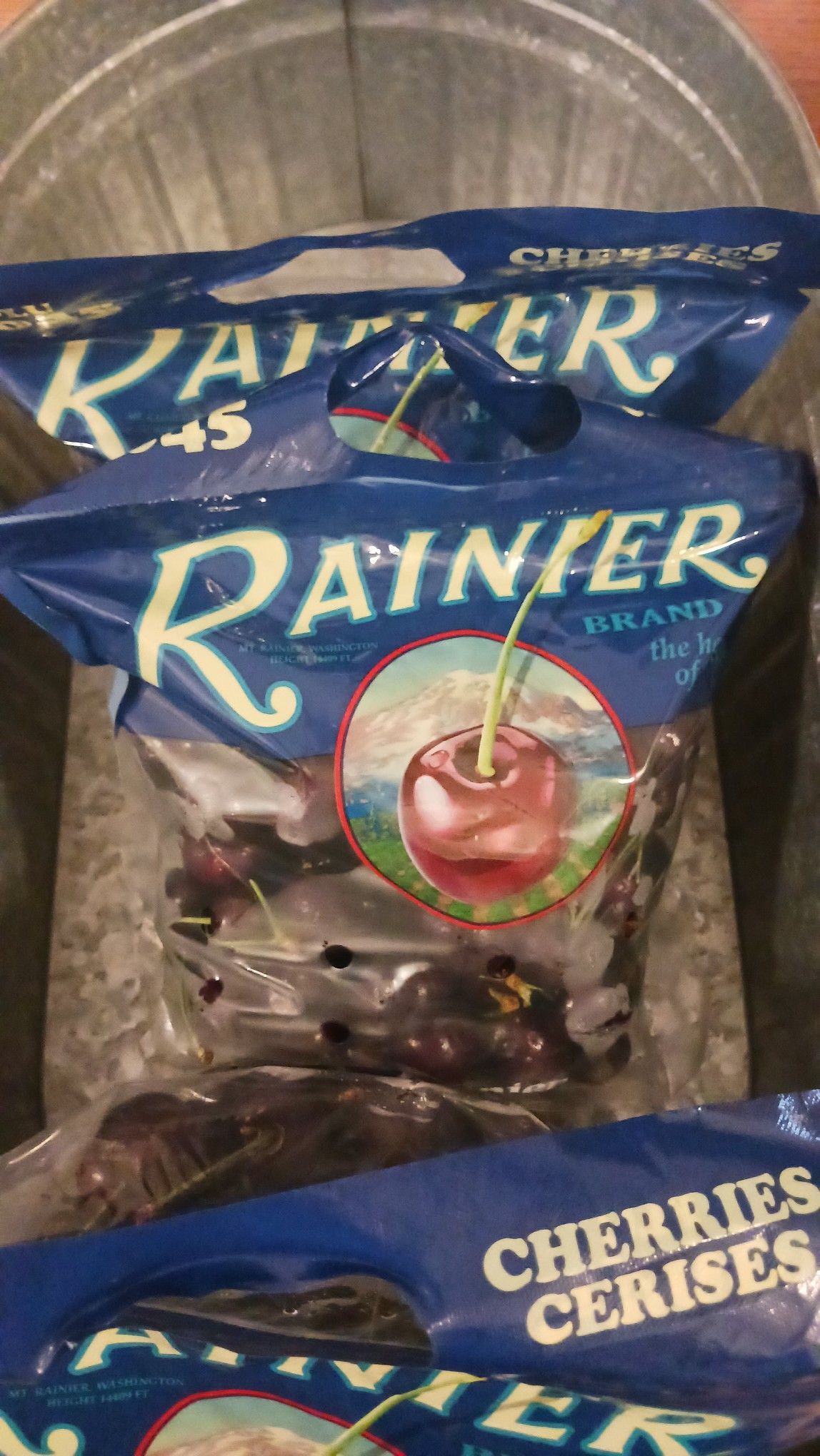 Bags of Rainier brand cherries, blue packaging with a cherry graphic, in a metal bin.