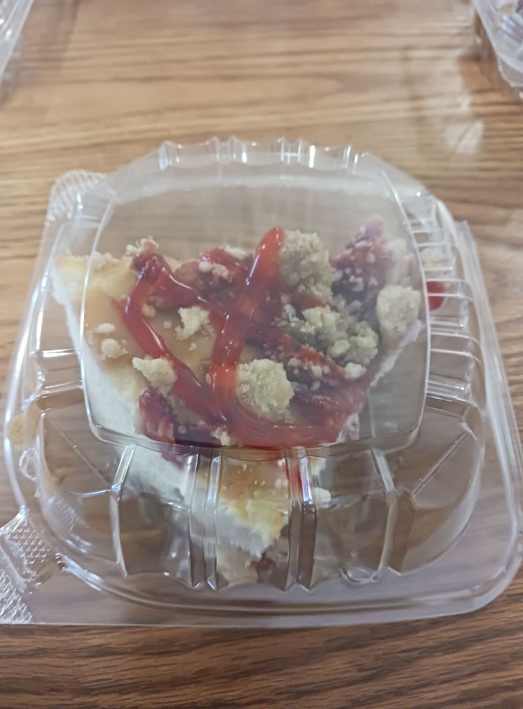 Slice of cheesecake with red topping in a clear plastic container on a wooden table.