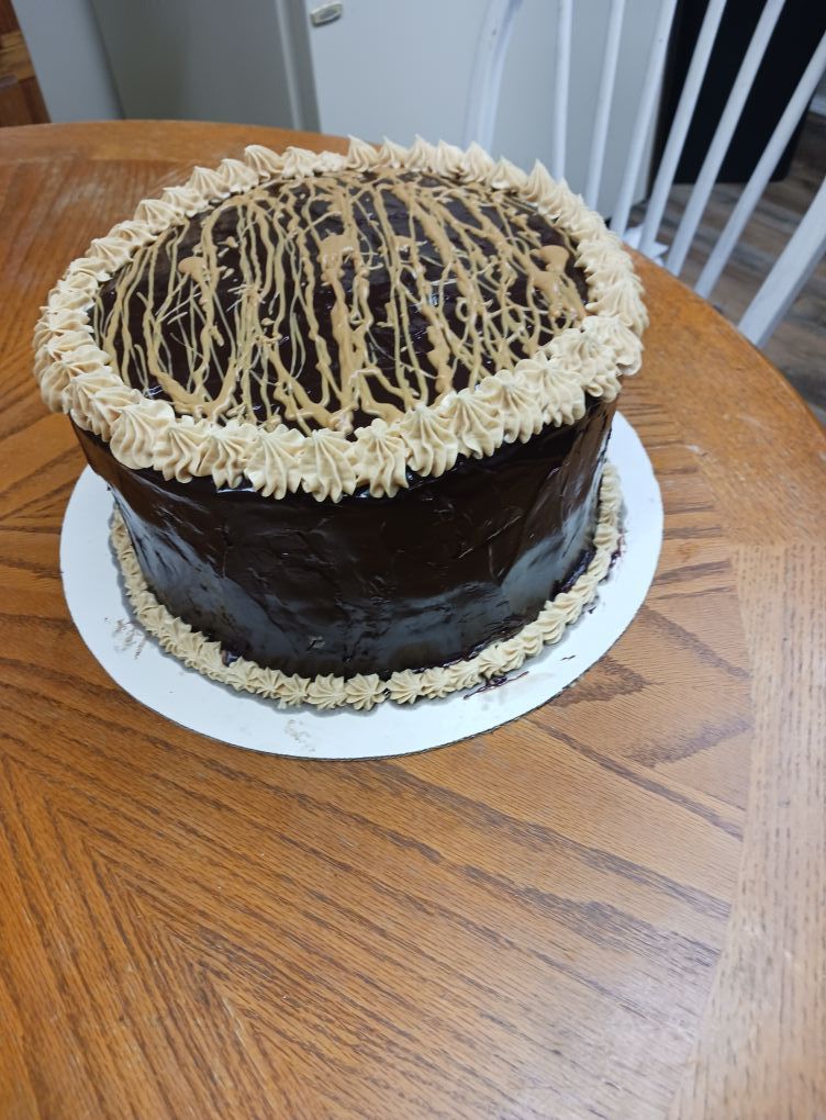 Chocolate cake on a white plate, with brown frosting and tan drizzle, set on a wooden table.