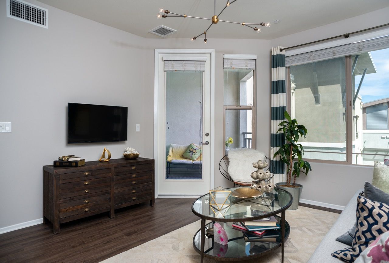 Living room in an apartment with a wall-mounted TV, wooden dresser, glass coffee table, and balcony door.