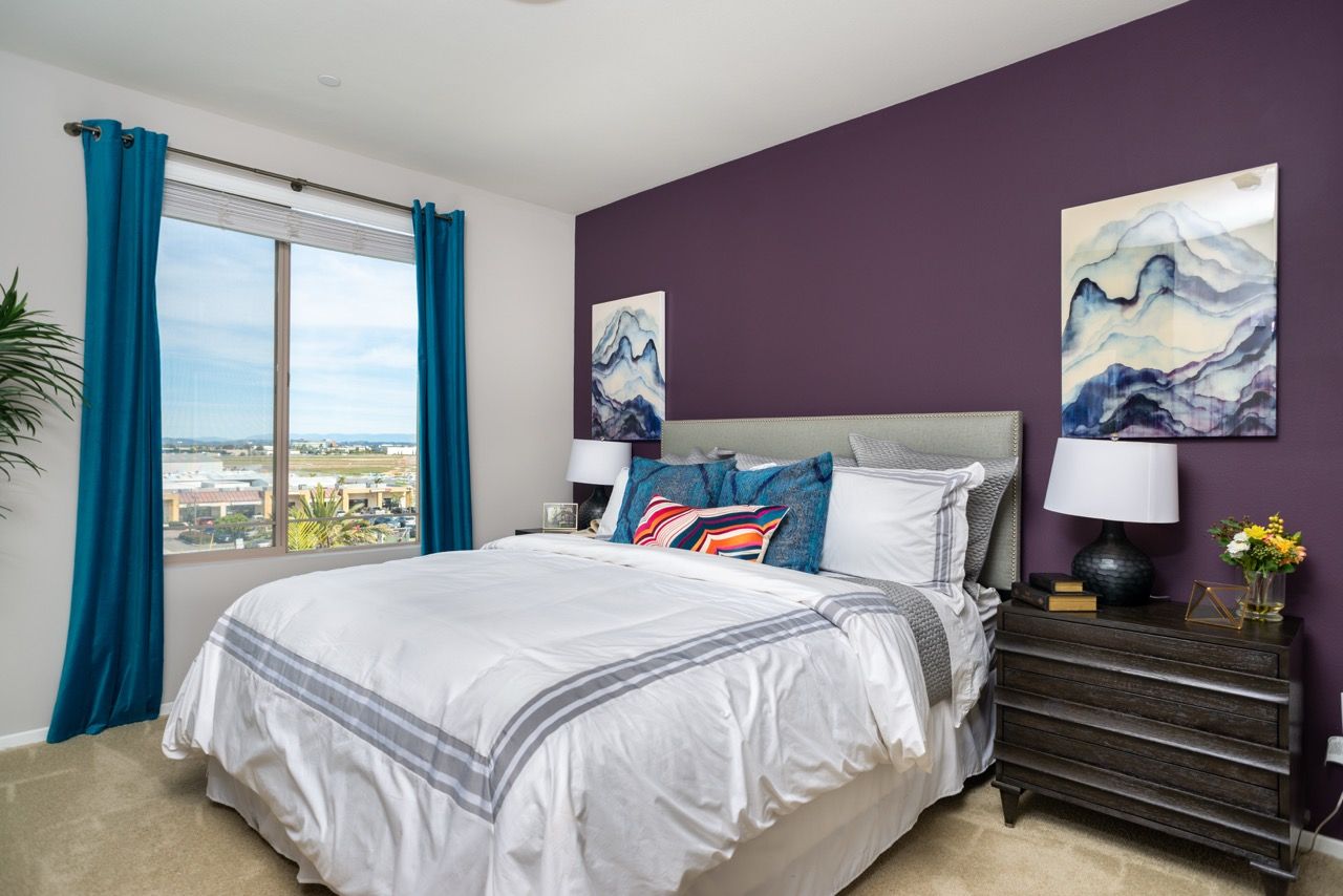 Bedroom with white bedding, purple accent wall, dark wood nightstand, and blue curtains.