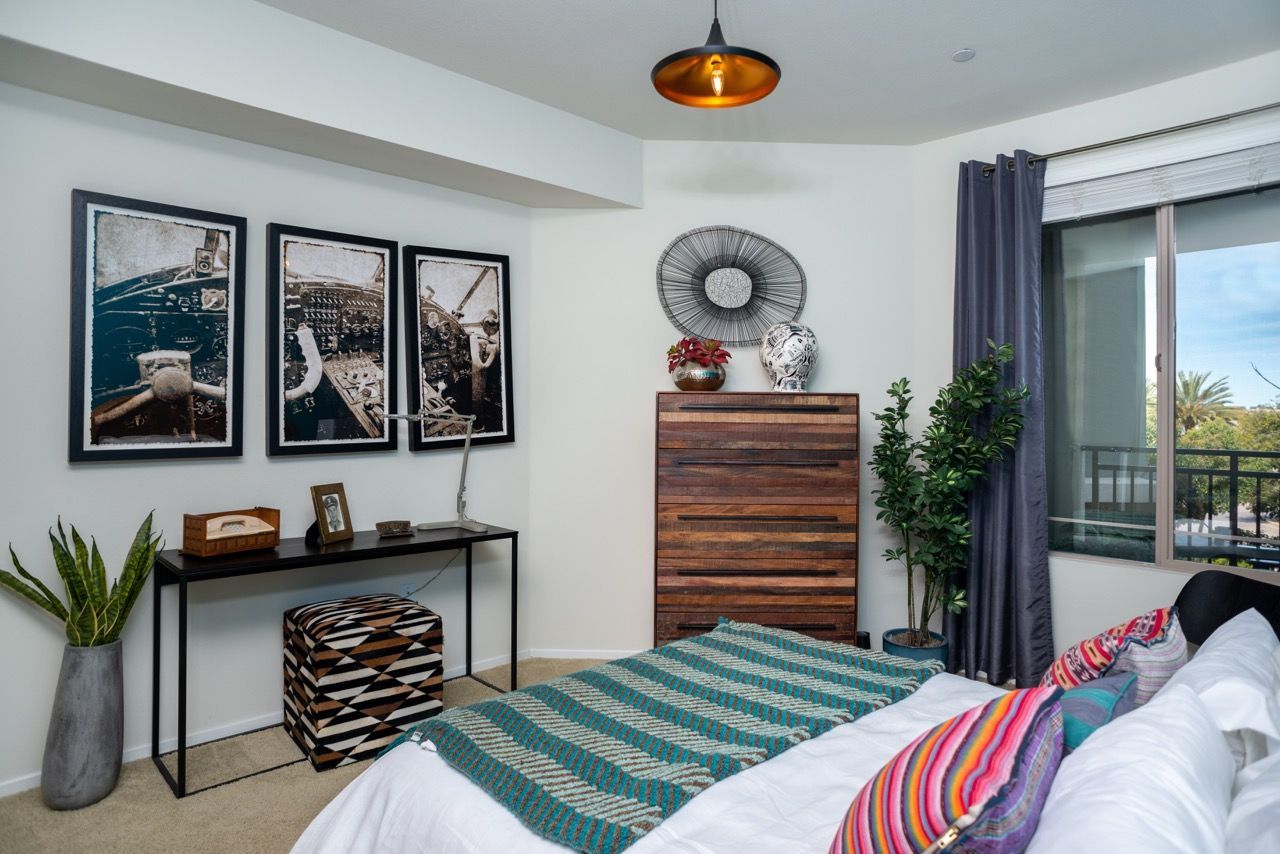 Bright bedroom in an apartment with a bed, dresser, plants, and wall art.