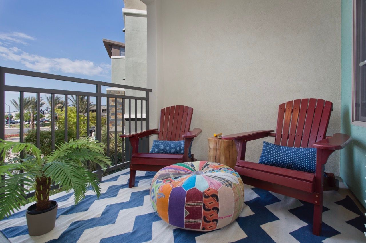 Balcony seating area with two red Adirondack chairs, a colorful pouf, and a potted plant on a patterned rug.