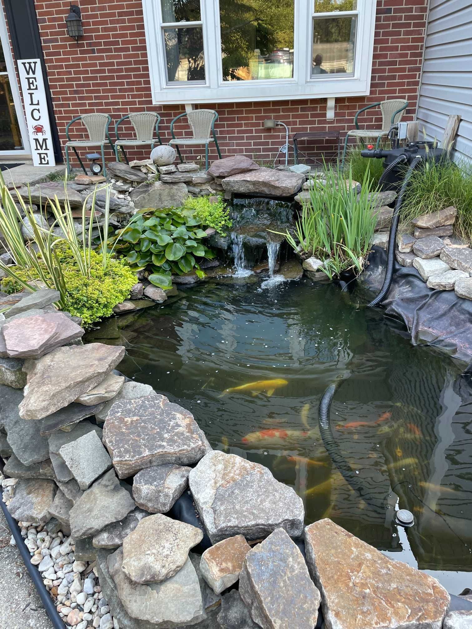 A backyard pond with waterfalls and several orange koi fish, surrounded by rocks and greenery.