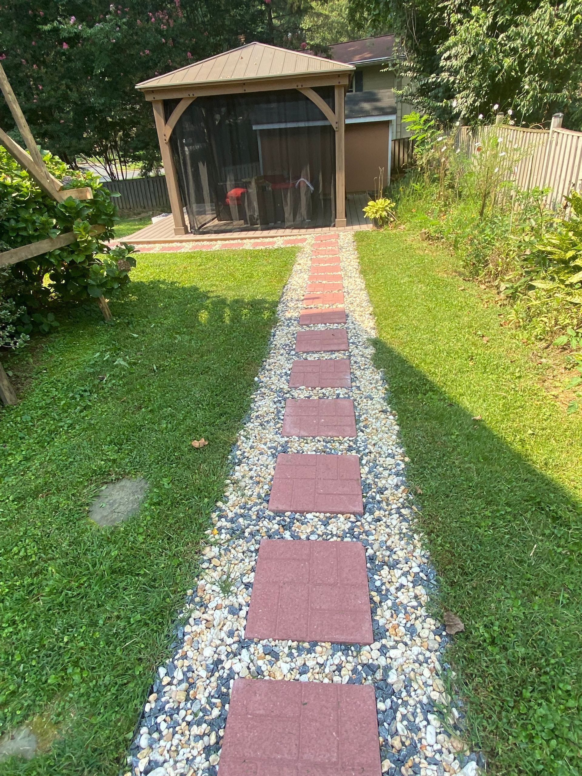 A stone pathway with red square pavers leading through a grassy yard toward a wooden gazebo.