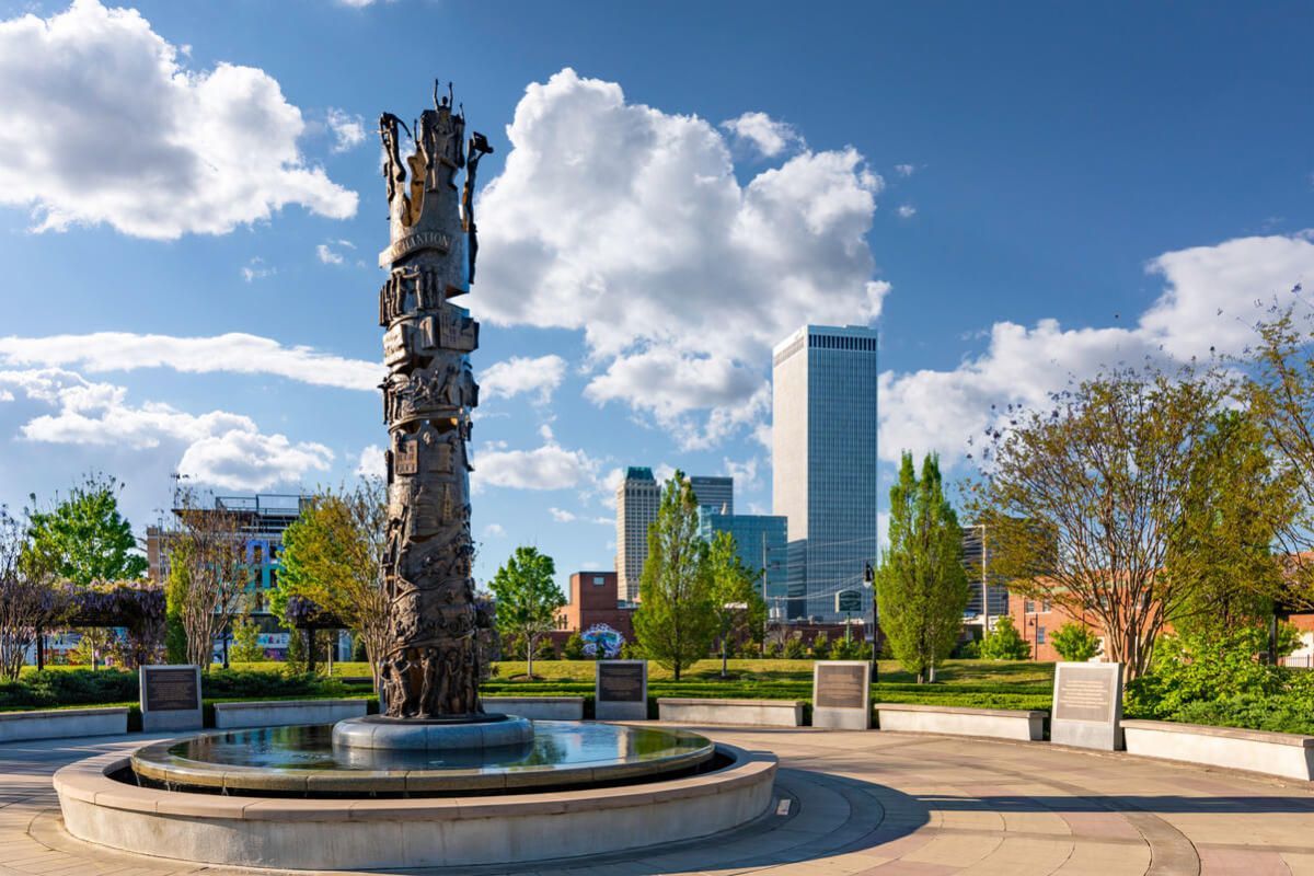 A bronze sculpture rises from a fountain in a park, with the Tulsa skyline visible against a blue, cloudy sky.