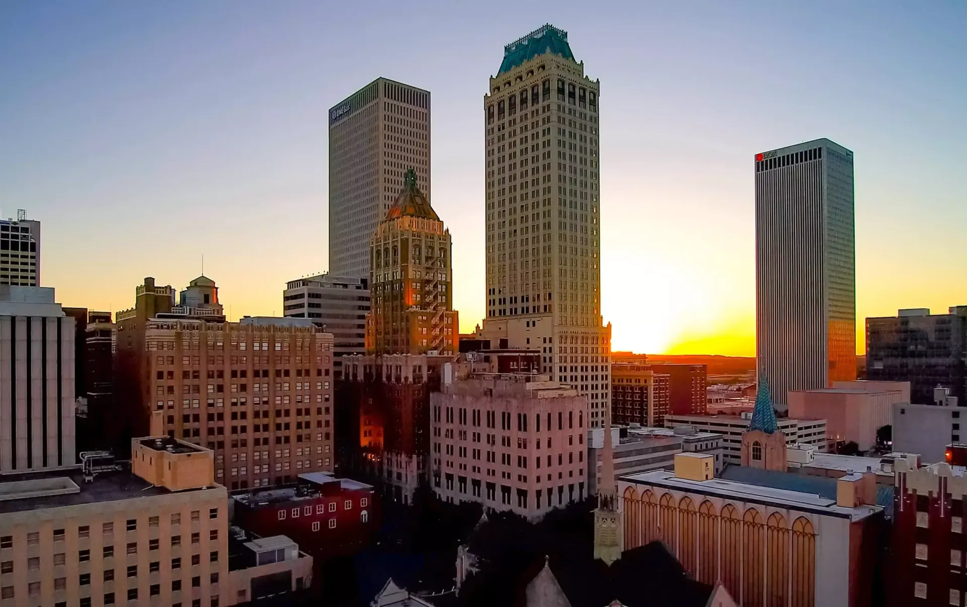 Sunset over the downtown Tulsa, Oklahoma skyline, featuring Art Deco skyscrapers and surrounding city buildings.