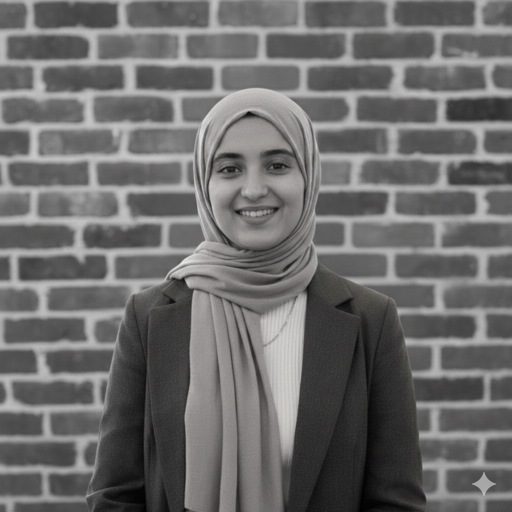 Woman wearing a hijab and blazer smiling in front of a brick wall.