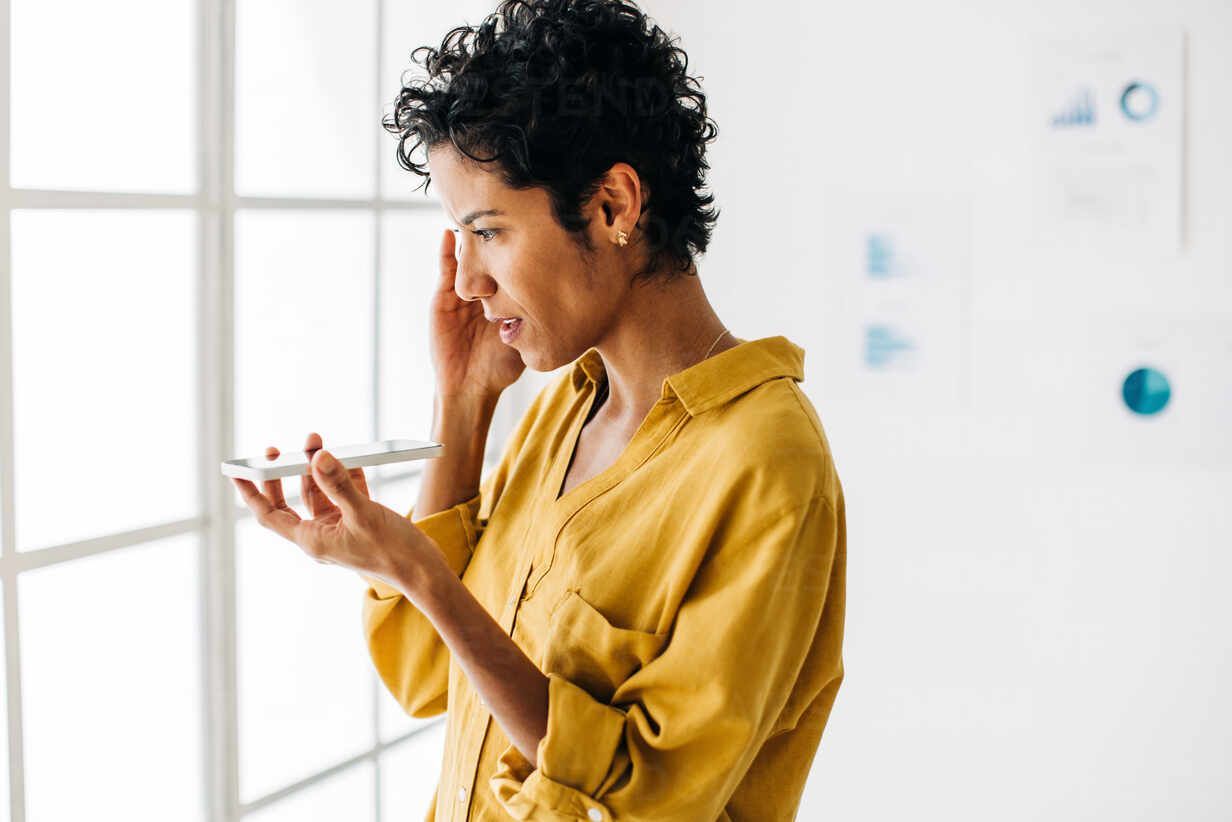 Woman in yellow shirt, holding a phone, near a window, talking, and looking intently.