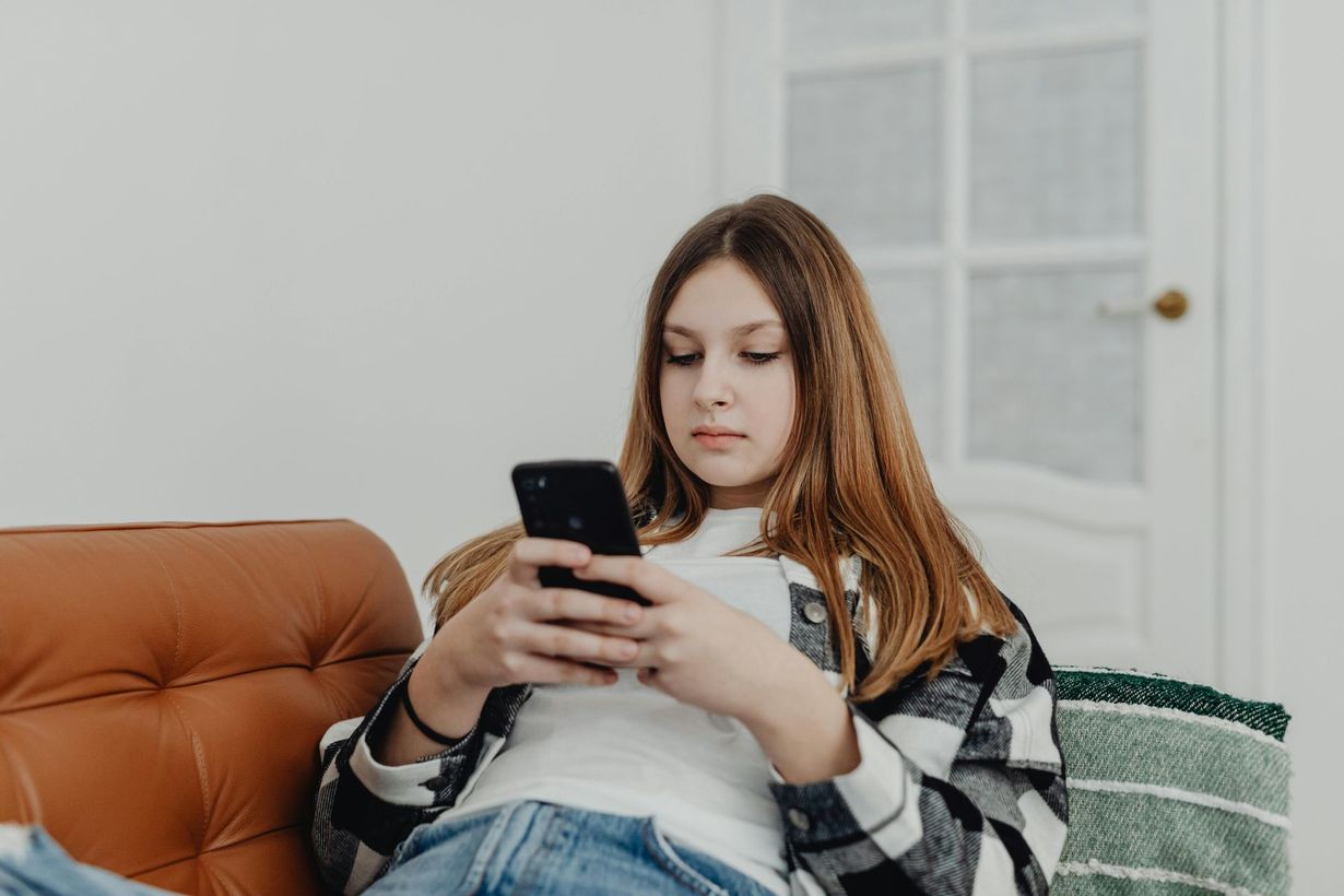 A person sits on a brown leather couch, looking down at a smartphone held in both hands.