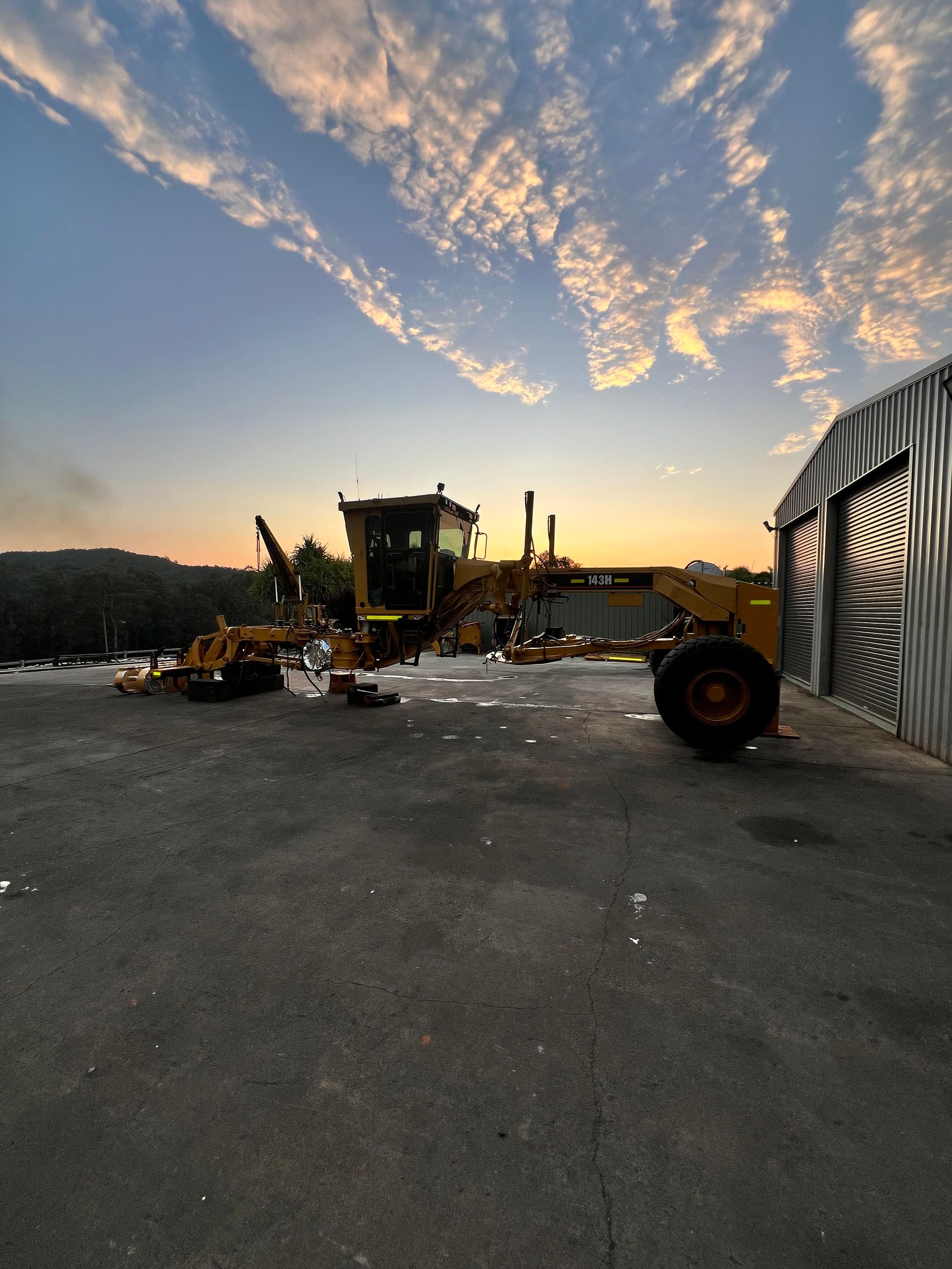 Road Grader Parked Outside a Corrugated Metal Building at Dusk, Orange Sky — HOLESHOT Diesel In Bundaberg, QLD