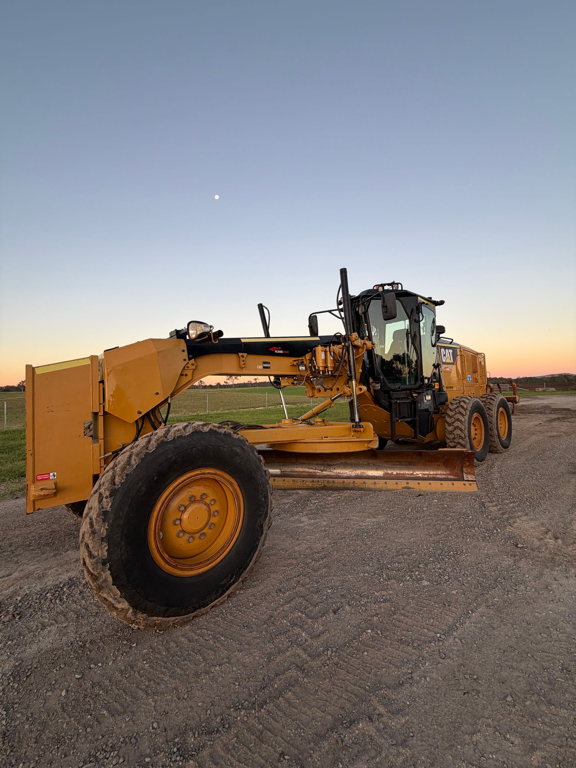 Yellow Motor Grader on Gravel in Front of a Blue and Orange Sky — HOLESHOT Diesel In Kingaroy, QLD