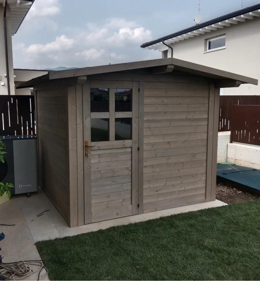 Capanno in legno con porta e finestra, situato su un prato accanto a una casa.