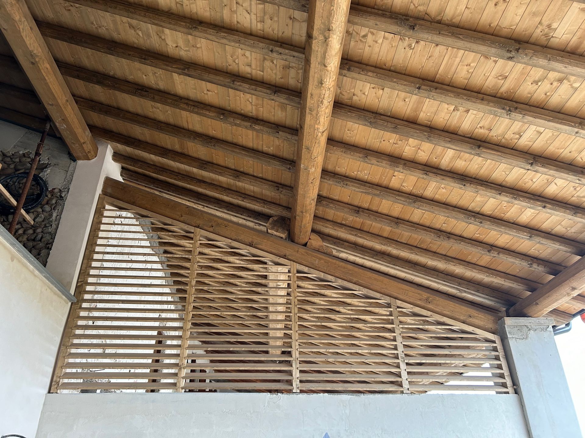 Tenda da sole con doghe in legno sopra un portico con travi di sostegno diagonali, soffitto in legno naturale e parete strutturata.