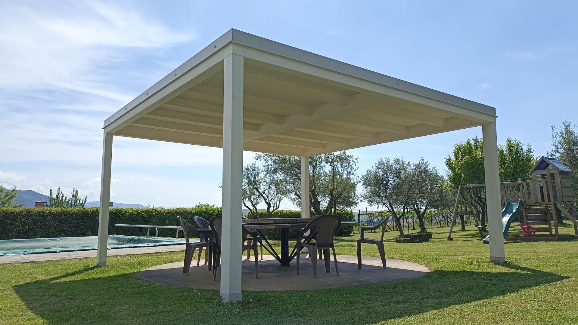 Pergolato con tavolo e sedie su un patio in un cortile erboso, alberi sullo sfondo.