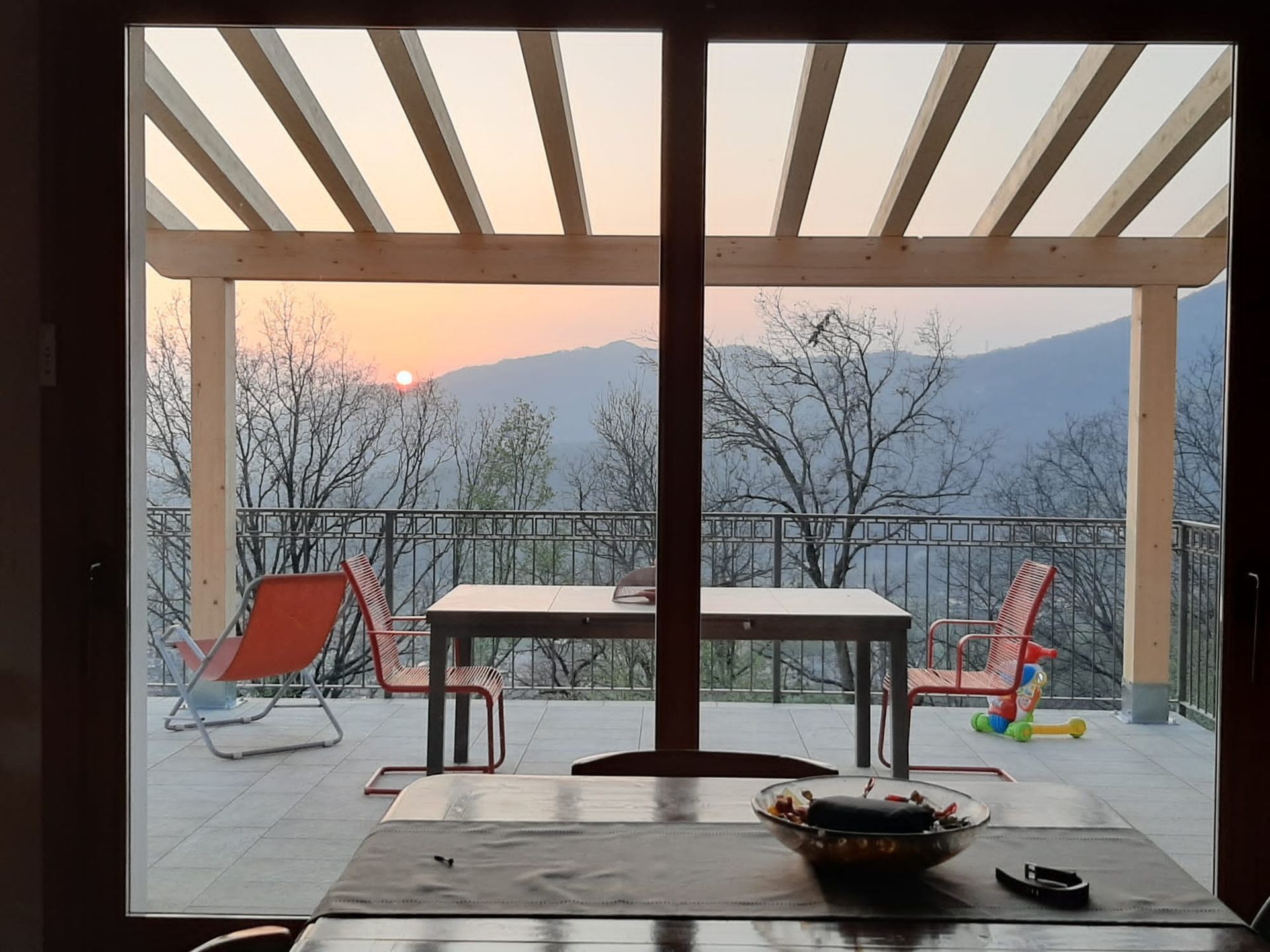 Vista attraverso una finestra di un tramonto sulle montagne, con pergolato e mobili da giardino.