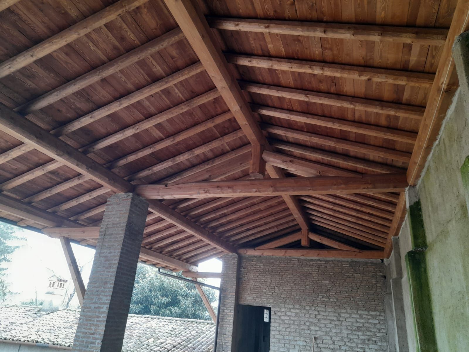 Soffitto in legno e travi sopra un portico sostenuto da pilastri in mattoni e muro in pietra.