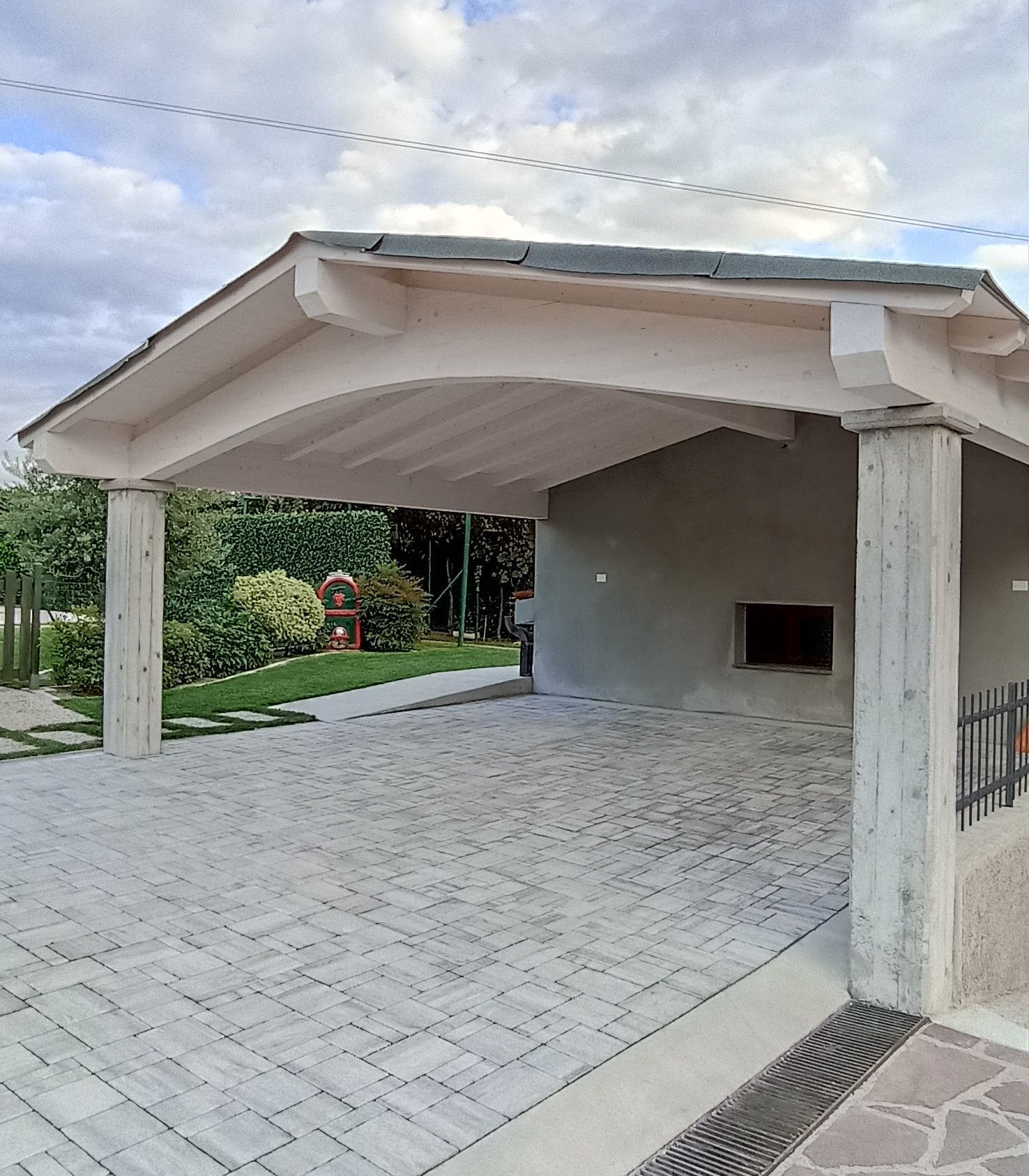 Carport con pavimentazione in mattoni grigi, sostenuto da colonne di cemento. Piante verdi e cielo nuvoloso sullo sfondo.